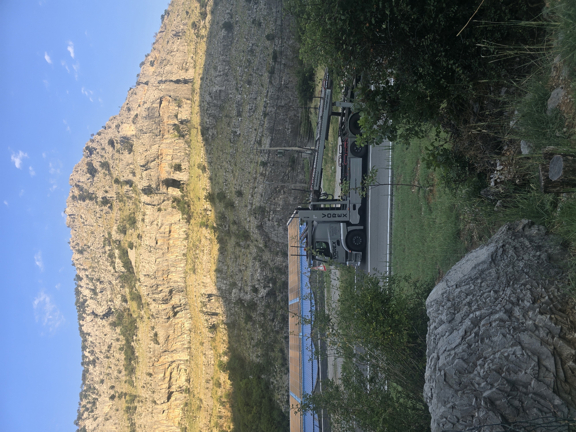 A large, grey truck with "VOREX" written on its side is driving on a highway. The truck is carrying some equipment on its trailer, which appears to be a car carrier. The highway is flanked by green grass and bushes. In the background, a large, rocky mountain rises into a clear blue sky. The mountain has a rough texture with visible strata and some sparse vegetation growing on its slopes. There is also a cave-like opening visible on the mountain face. Sunlight illuminates the mountain, casting shadows on its surface and highlighting its rugged contours. The scene appears to be in a rural or mountainous area.