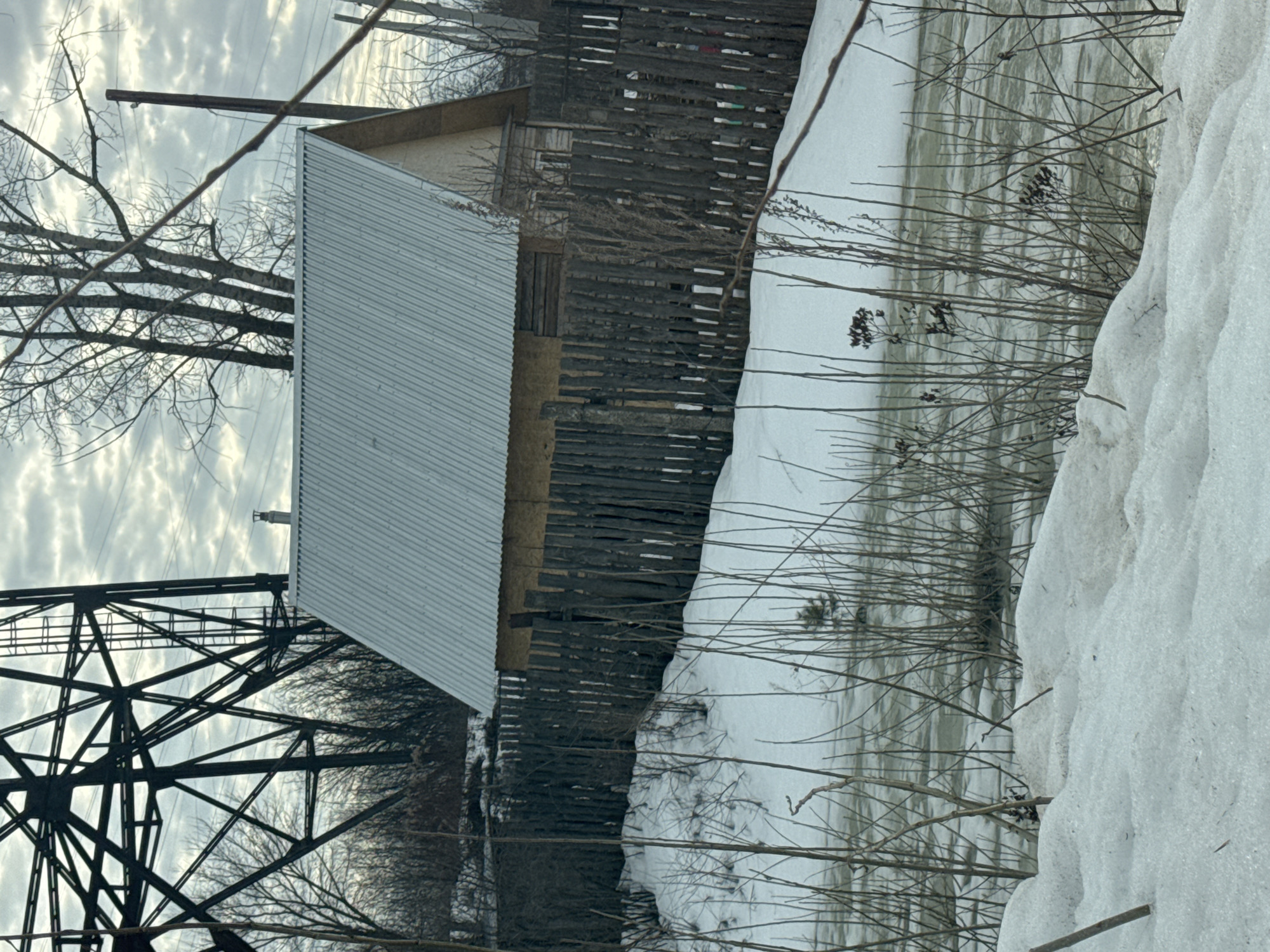 The image depicts a winter scene, potentially in Chaikovsky, Russia (although this cannot be definitively confirmed without more information). A metal-roofed structure is prominently featured, situated next to a weathered wooden fence. A bridge or powerline structure, composed of intersecting metal beams, is visible in the lower left corner. Snow covers the ground, with a significant accumulation along the right side of the frame. Bare branches of trees and bushes are visible throughout, adding to the wintry atmosphere. The sky is overcast. The overall impression is of a rural or semi-rural setting.