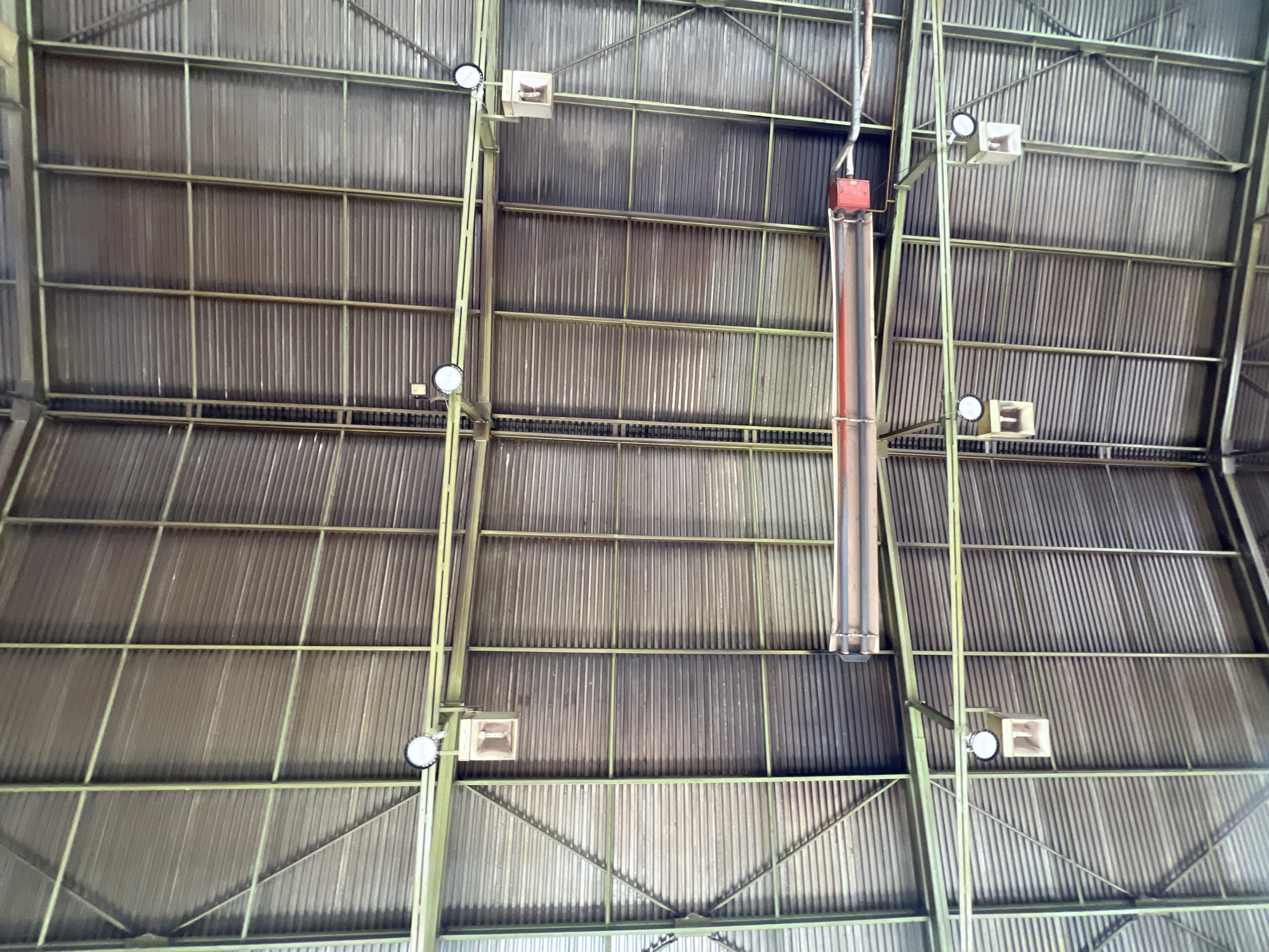 The image shows the interior of a large, industrial-style building, likely a gymnasium or a warehouse, looking directly up at the corrugated metal ceiling and the supporting metal framework. The ceiling is constructed with a series of parallel, ridged metal sheets, supported by a grid of light green painted metal beams and trusses.
Several light fixtures are visible, each consisting of a circular, white LED light attached to a mounting bracket, with a rectangular, light-colored diffuser box hanging below. These are positioned at intervals across the ceiling structure. In the central upper portion of the image, a long, reddish-brown cylindrical object, possibly a heating element or a pipe, is suspended horizontally, with a red control box attached to its right end.
The overall impression is one of a functional, utilitarian space. There are no people or animals visible in the image. The lighting suggests it is daytime, with the natural light likely filtering in from unseen sources, though artificial lights are also present. The building's structure is the primary focus, with the metal framework and the ribbed ceiling dominating the visual field. The image has been taken from a low angle, looking upwards. The location is identified as Lanžhot, Czechia, suggesting this is a structure within that city.