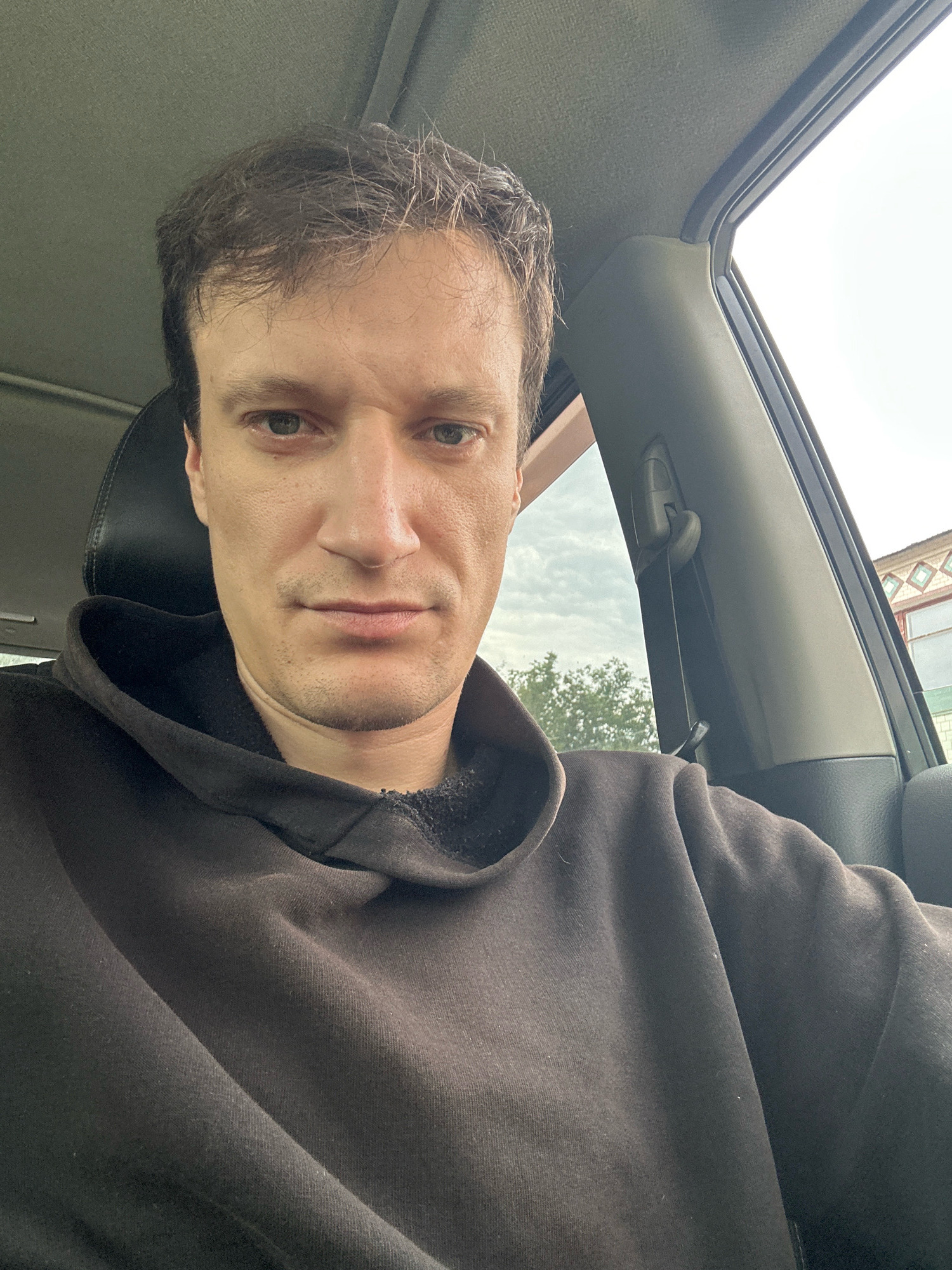 A man with short, brown hair is sitting in the driver's seat of a car, looking directly at the camera. He is wearing a dark brown hooded sweatshirt. The interior of the car is visible, including a black seat and part of the car's roof lining. The window on the right shows an overcast sky and trees. A building with a distinctive roofline is visible in the background through the window. The scene appears to be located in Makiv, Ukraine.