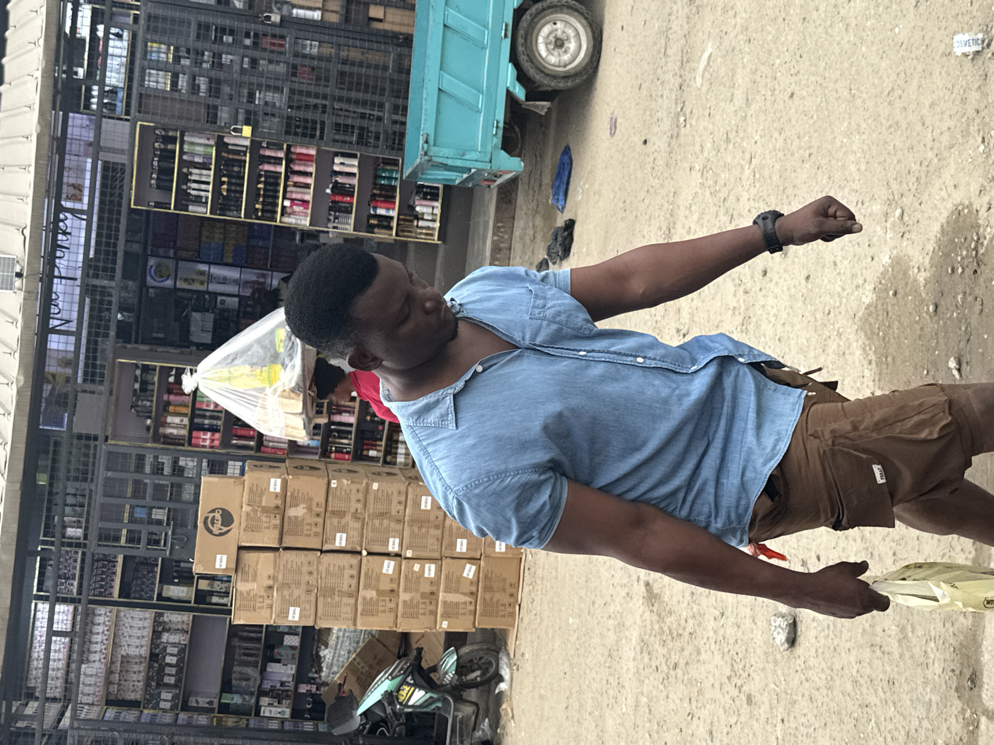 A man wearing a blue denim shirt and brown cargo shorts walks in the foreground of an outdoor market in Satellite Town, Nigeria. He is carrying a light-colored plastic bag in his left hand and has a dark-colored watch on his left wrist. Behind him, shelves stocked with various products, including what appear to be cosmetics and toiletries, are visible. A turquoise truck is parked to the right. A light green and black scooter is parked to the left. The ground is a mix of sand and dirt.