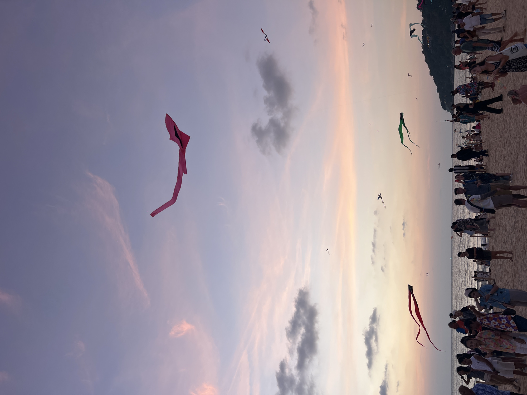 The image shows a beach scene at sunset, likely in Karon, Thailand. The sky is a mix of lavender, pink, and orange hues with scattered clouds. Several kites are flying in the sky, including a prominent pink kite on the left side of the frame, a green kite in the center, and a red kite near the bottom of the image. The beach is crowded with people, many of whom appear to be watching or photographing the kites. The image appears to be taken on a beach, with a large crowd of people present. A slight hill or embankment can be seen beyond the beach.