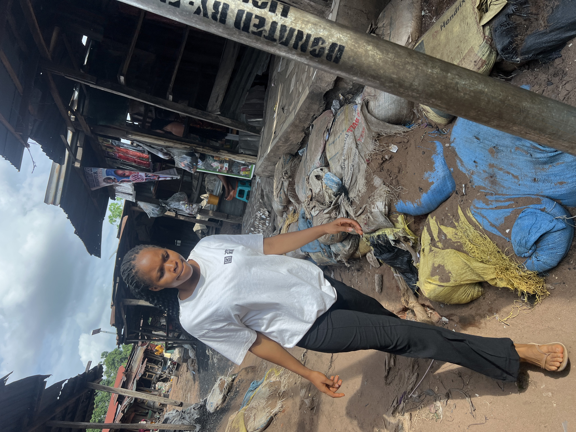 A young woman in a white t-shirt and black pants stands barefoot in a cluttered, potentially hazardous area of Unknown, Unknown. The environment includes worn structures made of wood and corrugated metal, and discarded sacks of various colors (blue, yellow, gray) are scattered on the ground, which appears to be dirt. A metal pole with lettering is visible in the foreground. The sky is overcast.