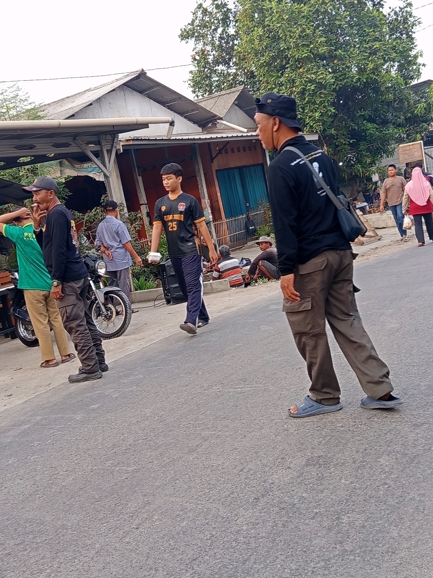 The image depicts a street scene in Karawang, Indonesia, with several individuals present. In the foreground, a man wearing a black long-sleeved shirt, cargo pants, a black hat, and blue sandals is walking away from the camera. He has a dark bag slung over his shoulder. To his left, another man in a black long-sleeved shirt, a black cap, and beige pants is smoking a cigarette and standing next to a motorcycle. Further to the left, a person in a green t-shirt and beige pants is leaning against the motorcycle. In the middle ground, a young man in a black and yellow jersey with the number "25" is walking, carrying a white object. Behind him, a man in a blue shirt and pants is walking, and further back, a woman in a pink hijab is seen. Several other individuals are seated or standing in the background near buildings. The environment appears to be a residential or commercial area with a paved road, some overhead structures, and trees.