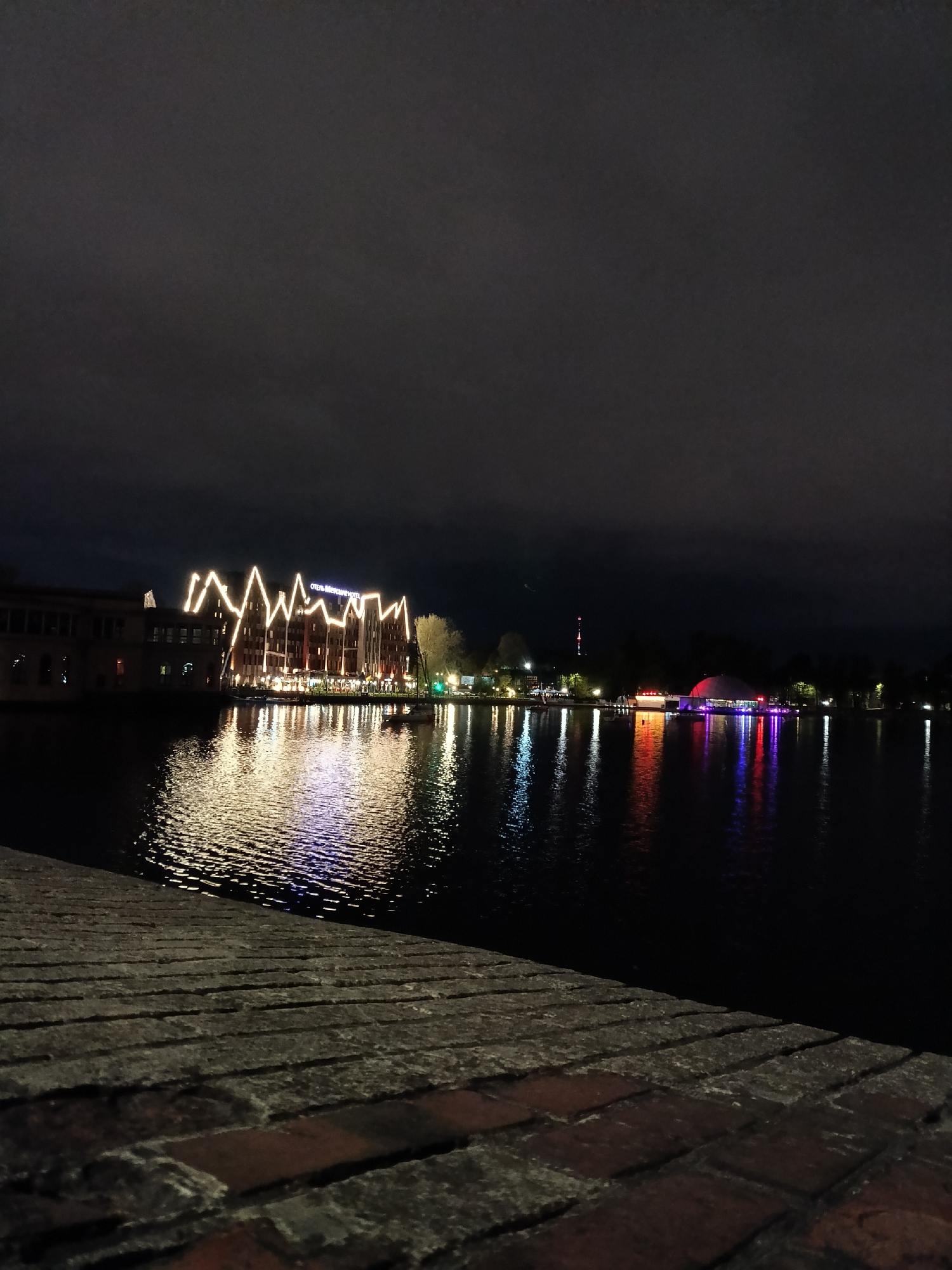 The image depicts a nighttime scene in Kaliningrad, Russia, likely near the Pregolya River or a connected body of water. A brick embankment is visible in the foreground, providing a ground-level perspective. Across the water, there's a building illuminated with white lights in a zig-zag pattern along its roofline, possibly the "Отель Микояновский Hotel," and lights reflecting in the water create a shimmering effect. Further in the distance, colored lights suggest the presence of other structures, possibly restaurants, bars, or a recreational area, with their reflections also visible in the water. The sky above is dark with cloud cover. There are no people visible in the image.