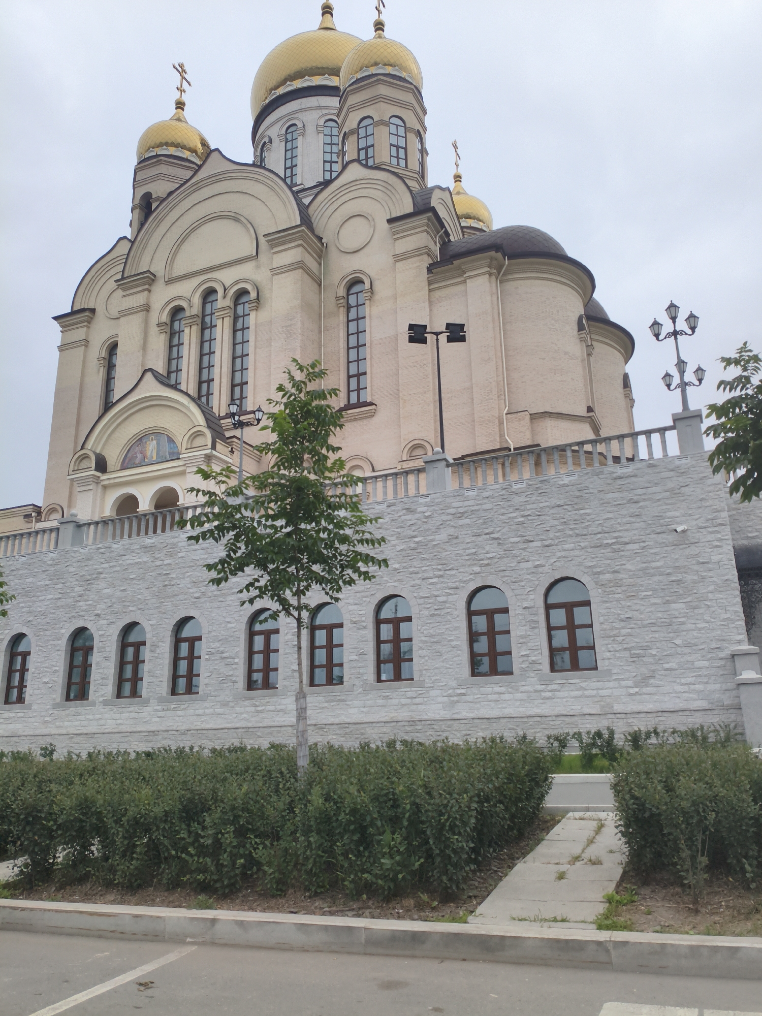 The image depicts a large, multi-domed religious building, likely a church, with ornate architecture. The building is primarily light-colored stone or stucco and features prominent golden domes topped with crosses. There are multiple rows of arched windows with dark frames on the lower section of the building, which is faced with lighter colored stone. A low wall with a balustrade runs along the front of the elevated section. A small tree stands in front of the building, and a path leads towards it between manicured bushes. A lamppost with multiple lights is visible on the right side of the frame, and another modern light fixture is seen on the left. The sky is overcast. This is the Church of the Archangel Michael in Vladivostok, Russia.