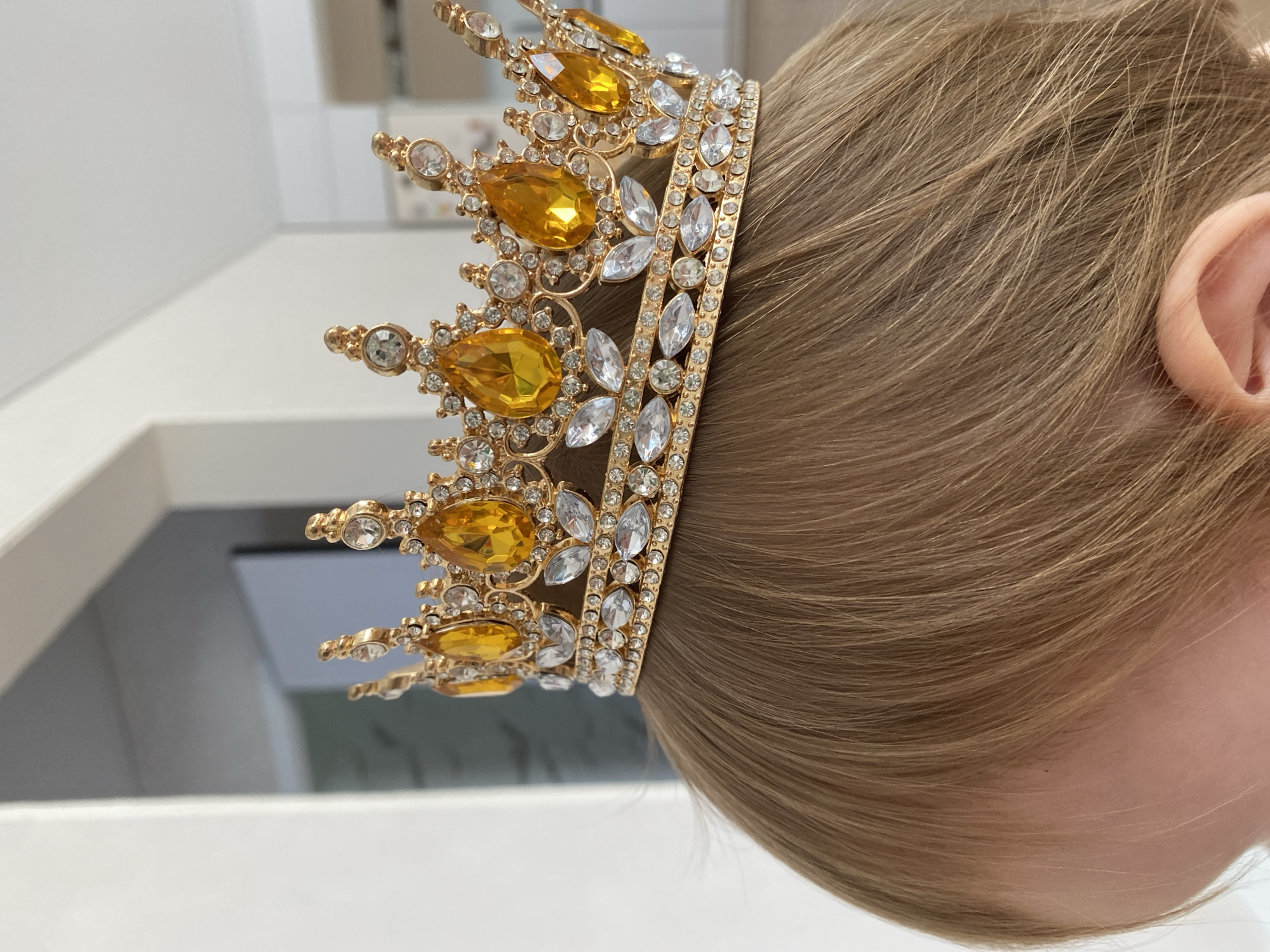 A close-up shot focuses on a golden crown adorned with yellow and clear rhinestones, placed on the head of a child with light brown hair. The crown features teardrop-shaped yellow stones and smaller clear stones arranged in decorative patterns. The child's hair is visible around the base of the crown, and a portion of their ear and cheek is also in view. The background is blurred, showing a bright, undefined indoor environment with hints of white walls and doorways. The lighting is bright and even, illuminating the details of the crown.