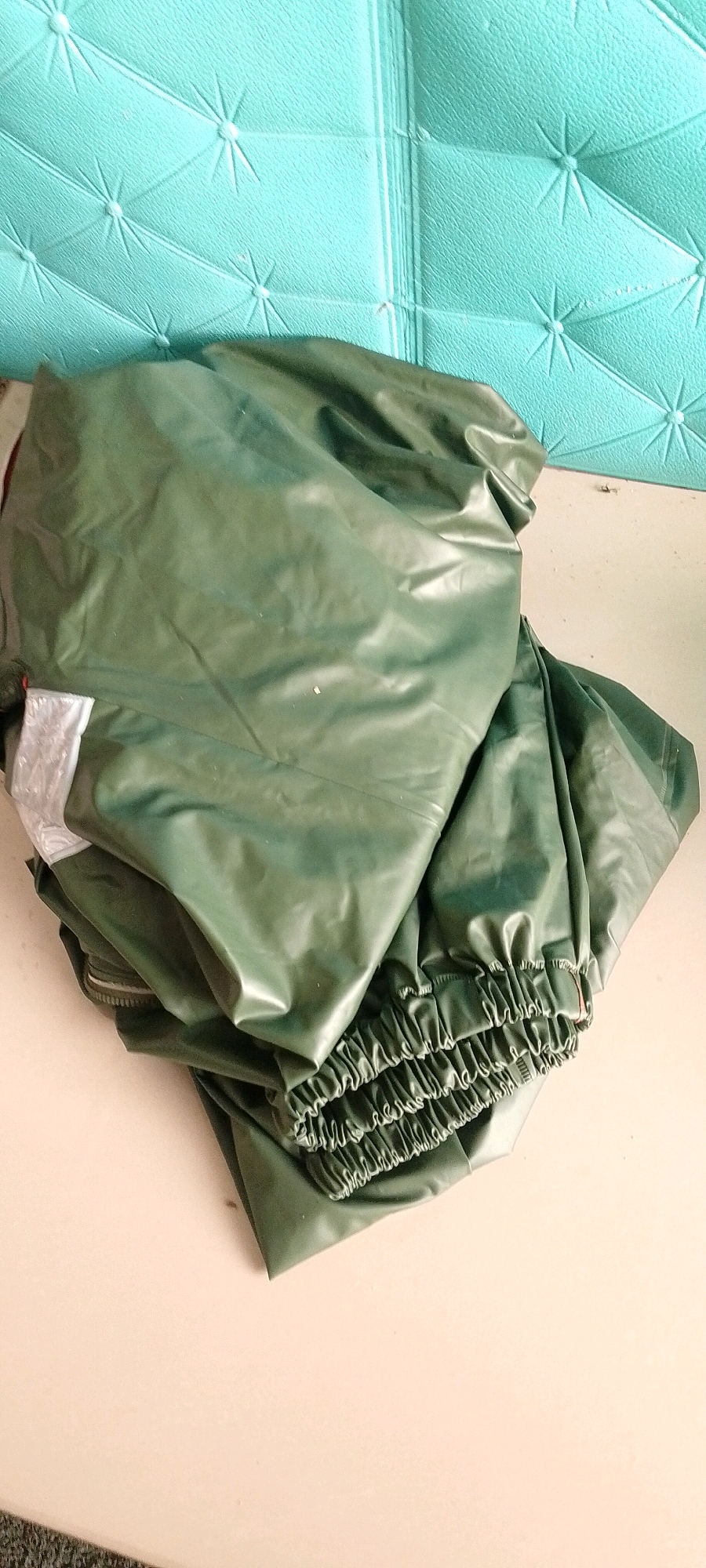 A pile of green fabric, possibly rain gear, is neatly folded on a light-colored surface. The fabric has a slightly shiny texture and exhibits wrinkles and folds, suggesting it is made of a pliable material like polyester or nylon. The upper portion of the image shows a textured turquoise background, possibly a wall or a decorative panel. The scene appears to be indoors, on a flat surface like a table or shelf. There are no people or animals visible in the image. The lighting suggests an ambient indoor light source. The image's composition places the folded fabric prominently in the foreground and middle ground, with the turquoise background filling the top portion. The overall impression is one of a neatly stored item.