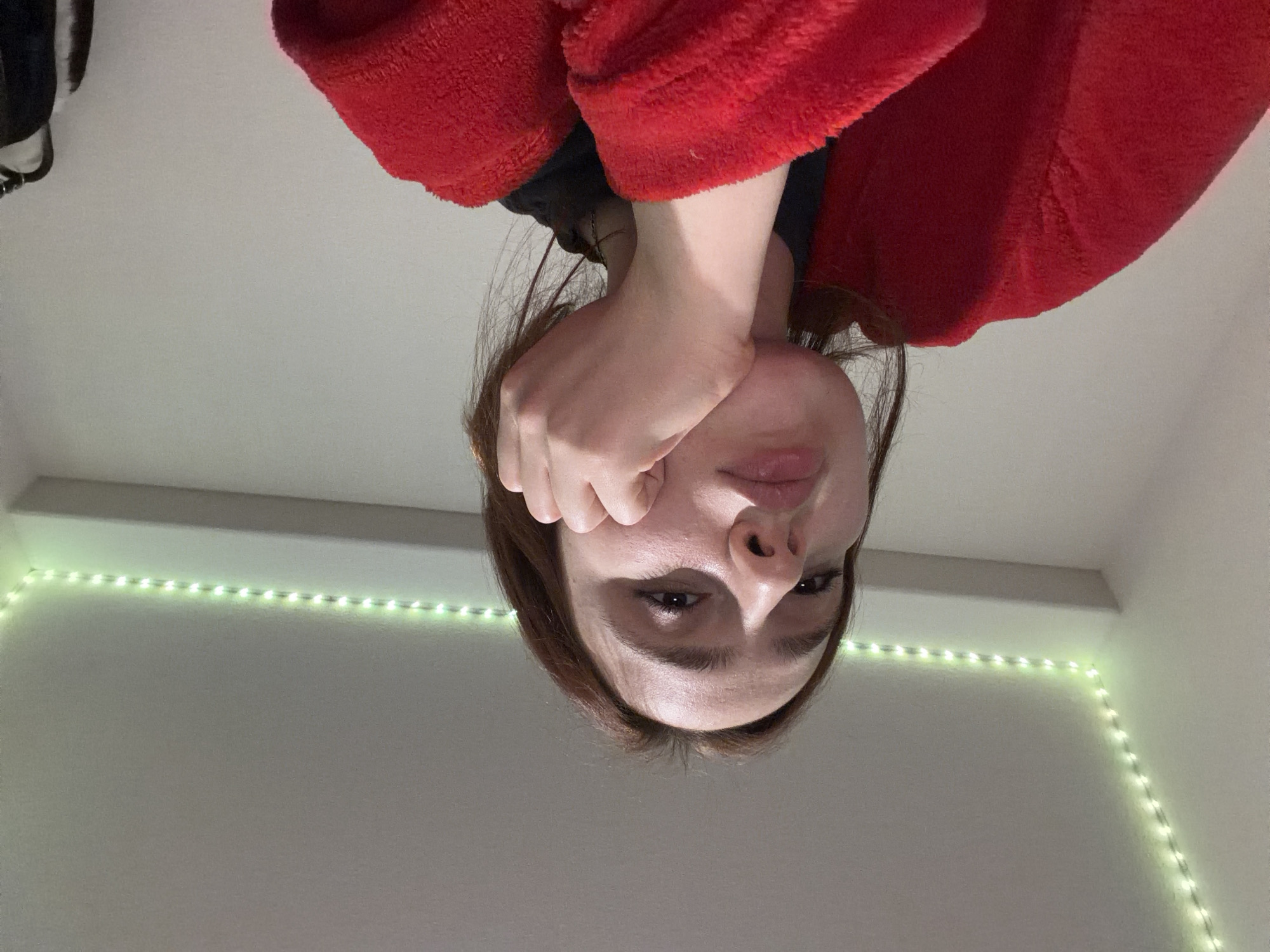 The image depicts a young female individual from a low-angle perspective, likely a selfie, in an indoor setting. The individual has light brown, shoulder-length hair and fair skin. Her eyes are directed slightly upward towards the camera, and her lips are slightly pursed. Her right hand is supporting her chin and lower jawline. She is wearing a plush, red long-sleeved garment, possibly a robe or fuzzy sweater, over a dark-colored top. The background consists of plain white or light-colored walls and a ceiling. Green LED strip lighting is installed along the top perimeter of the room, illuminating a corner where the wall meets the ceiling. A horizontal architectural feature, possibly a shelf or beam, is visible below the LED strip. A dark, indistinct object hangs partially visible on the wall to the far right. The specific location in the city of Strasbourg, France, is not determinable from the visual information provided.
