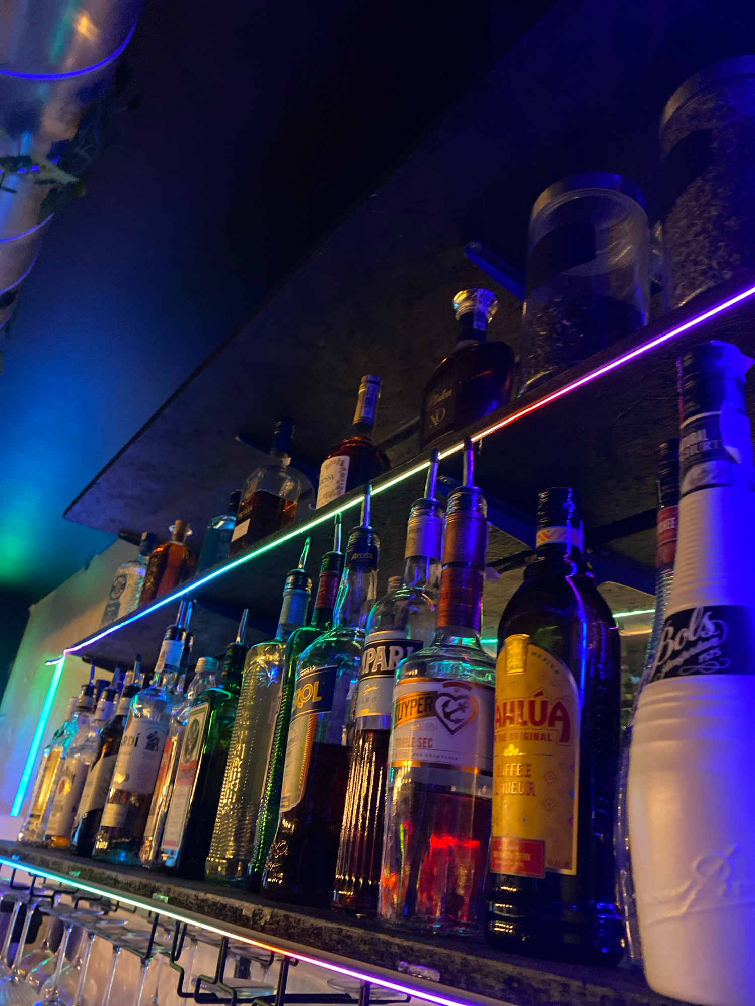The image displays a shelf of liquor bottles in a dimly lit establishment, possibly a bar. The bottles are of various shapes and sizes, with labels indicating different brands and types of alcohol. Some of the bottles are partially obscured by others. The shelf is illuminated by colorful LED strip lights, which cast a vibrant glow on the bottles and the surrounding environment. In the foreground, there are several wine glasses hanging upside down from a rack. The overall atmosphere is one of a nighttime establishment, possibly a club or bar. The image does not contain any information about the city of Gliwice, Poland, or any specific location.