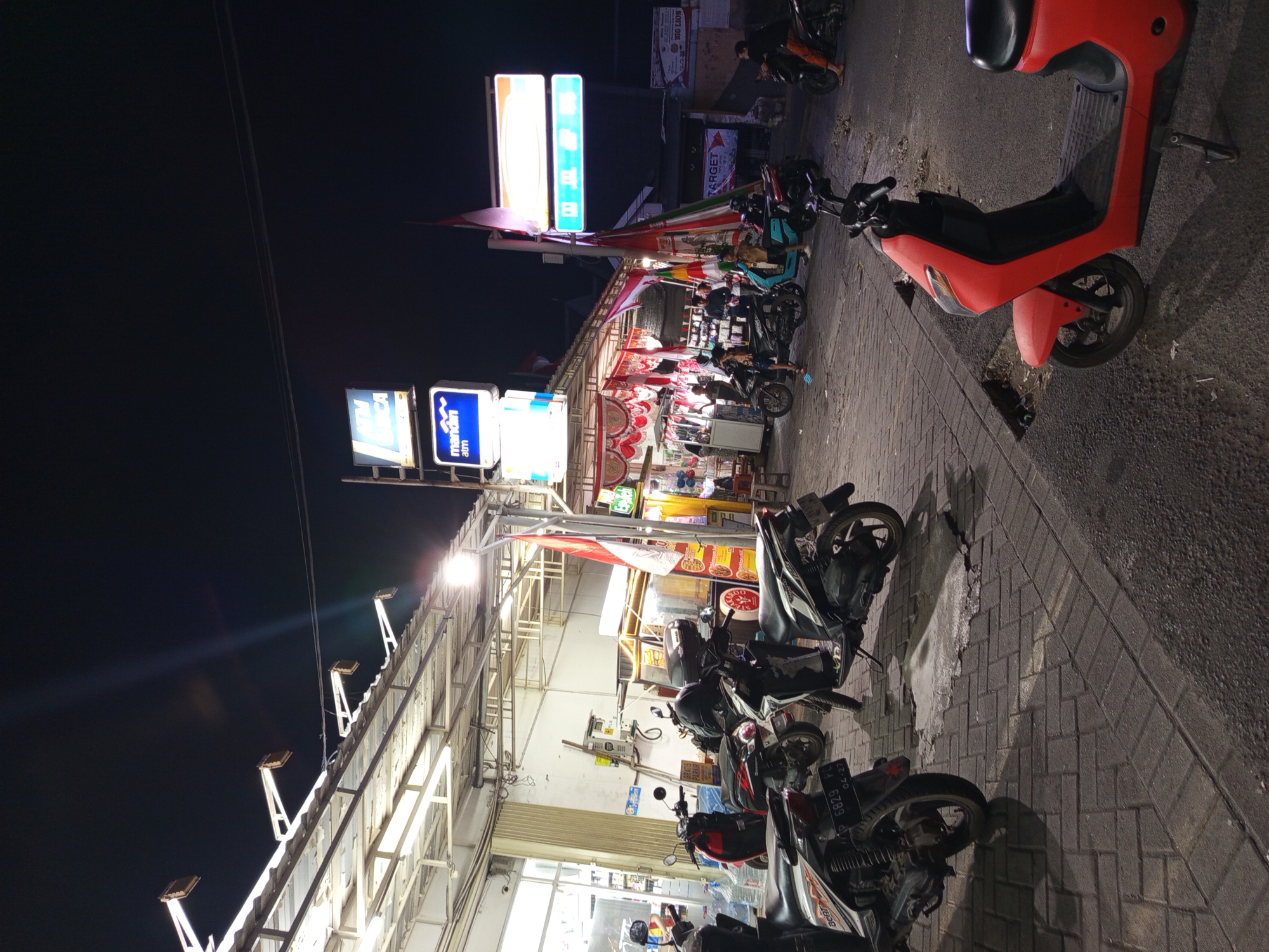 The image depicts a nighttime street scene in Gemurung, Indonesia, likely a commercial area. Several scooters and motorbikes are parked on a brick-paved sidewalk and the adjacent asphalt road. Overhead, a structure with fluorescent lighting and angled spotlights illuminates the area. Neon signs are visible, including one displaying "ATM MANDIRI" and another with Indonesian text. Flags, including the Indonesian national flag, are displayed. In the background, people are milling about near stalls and shops. The overall atmosphere suggests a busy marketplace or entertainment district after dark.