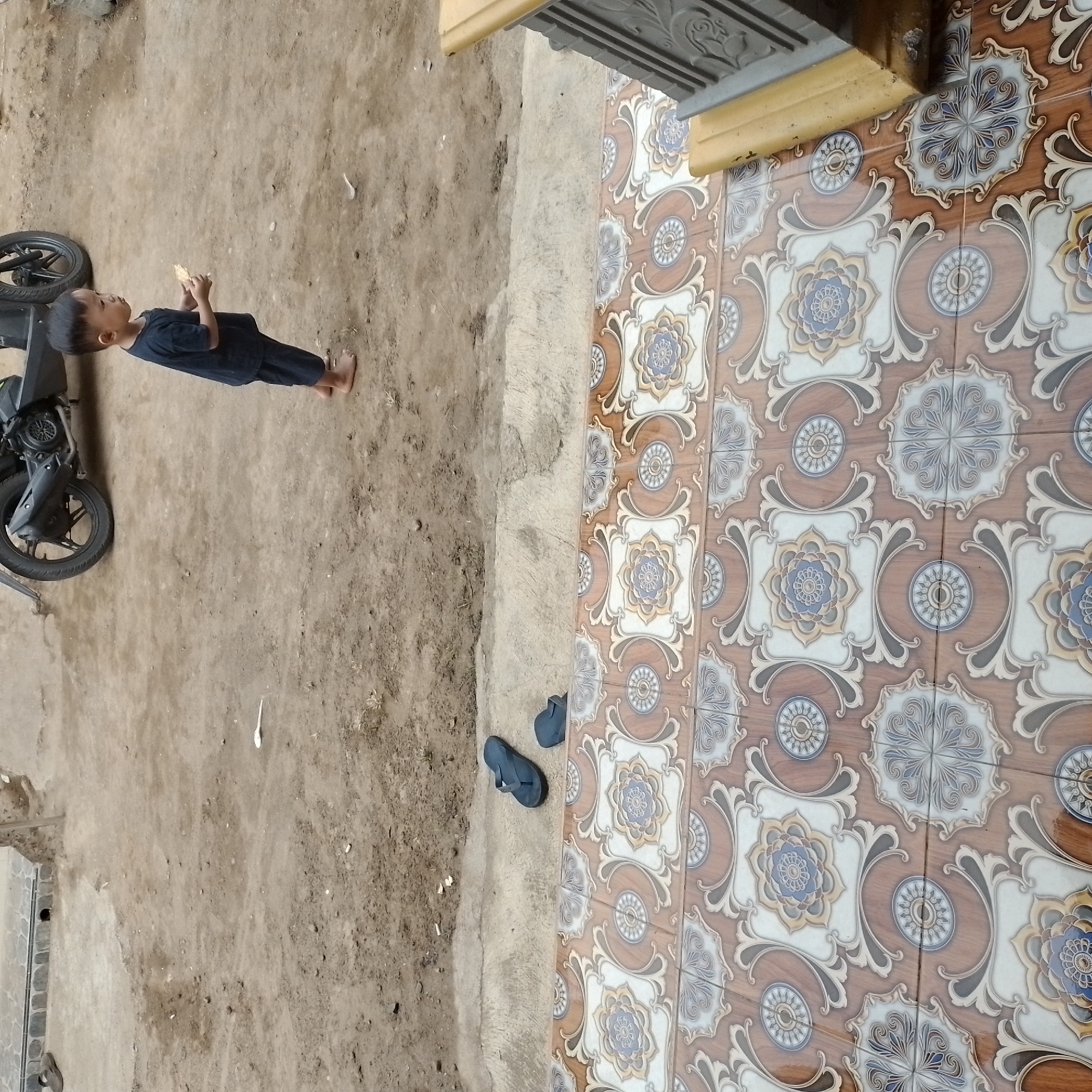 In Jatisobo, Indonesia, a young boy dressed in a dark blue t-shirt and shorts stands barefoot on a dirt ground. He holds a small, light-colored object in his hands, possibly a cracker or a piece of bread, and is looking at it. Behind him, a black motorcycle is parked. To the left of the boy, a pair of dark-colored flip-flops rests on the ground near a paved area. In the foreground, a patterned tiled floor with intricate floral designs in brown, white, and blue is visible. The environment appears to be an outdoor space, likely a yard or a pathway, with a mix of dirt and paved surfaces.