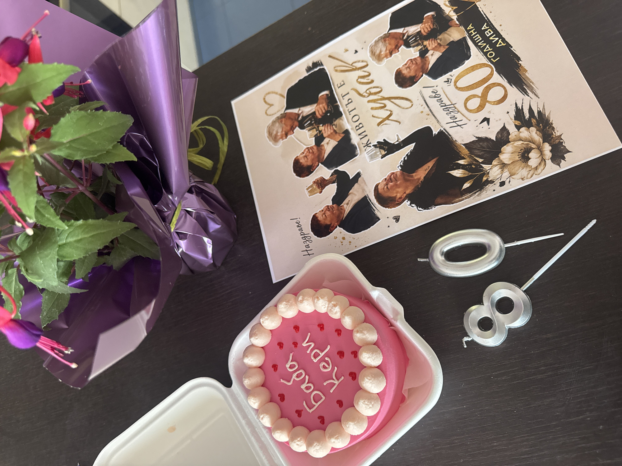 On a dark brown surface are several items arranged for a celebration, potentially a birthday. A bouquet of fuchsia flowers wrapped in purple cellophane is on the left. Next to it is a small square cake in a white container. The cake has pink frosting and is decorated with white icing dollops and tiny red hearts. Inscribed in white frosting on the cake are Cyrillic letters. Adjacent to the cake are silver number-shaped candles, forming "80." A card featuring several images of the same man, along with Cyrillic text and a floral design, completes the arrangement. Based on the Cyrillic text on the cake and card, this image is likely taken in a location where the Cyrillic alphabet is used, such as Burgas, Bulgaria.