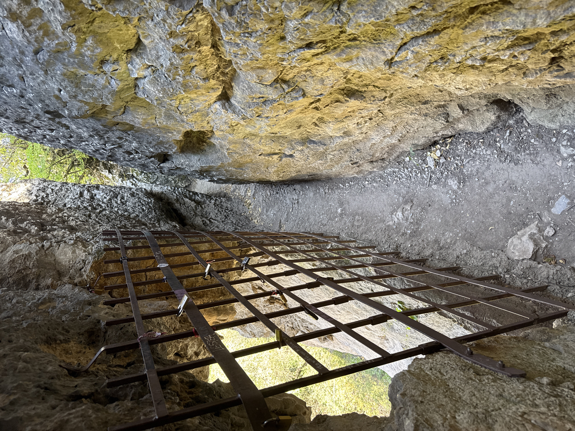 The image shows a narrow path between two rocky cliffs. On the left side, a metal grate separates the path from an opening in the rock face, which reveals a sunlit area with greenery outside. Several padlocks are attached to the grate, some with strings or ribbons. The path itself is composed of dirt and gravel, with loose stones and some fallen leaves. The rock walls are rough and textured, with crevices and natural formations. The lighting suggests it is daytime, with sunlight illuminating parts of the scene, particularly the right cliff face, creating strong shadows. This scene is located at the Hotnitsa Waterfall, in the village of Hotnitsa, Bulgaria.