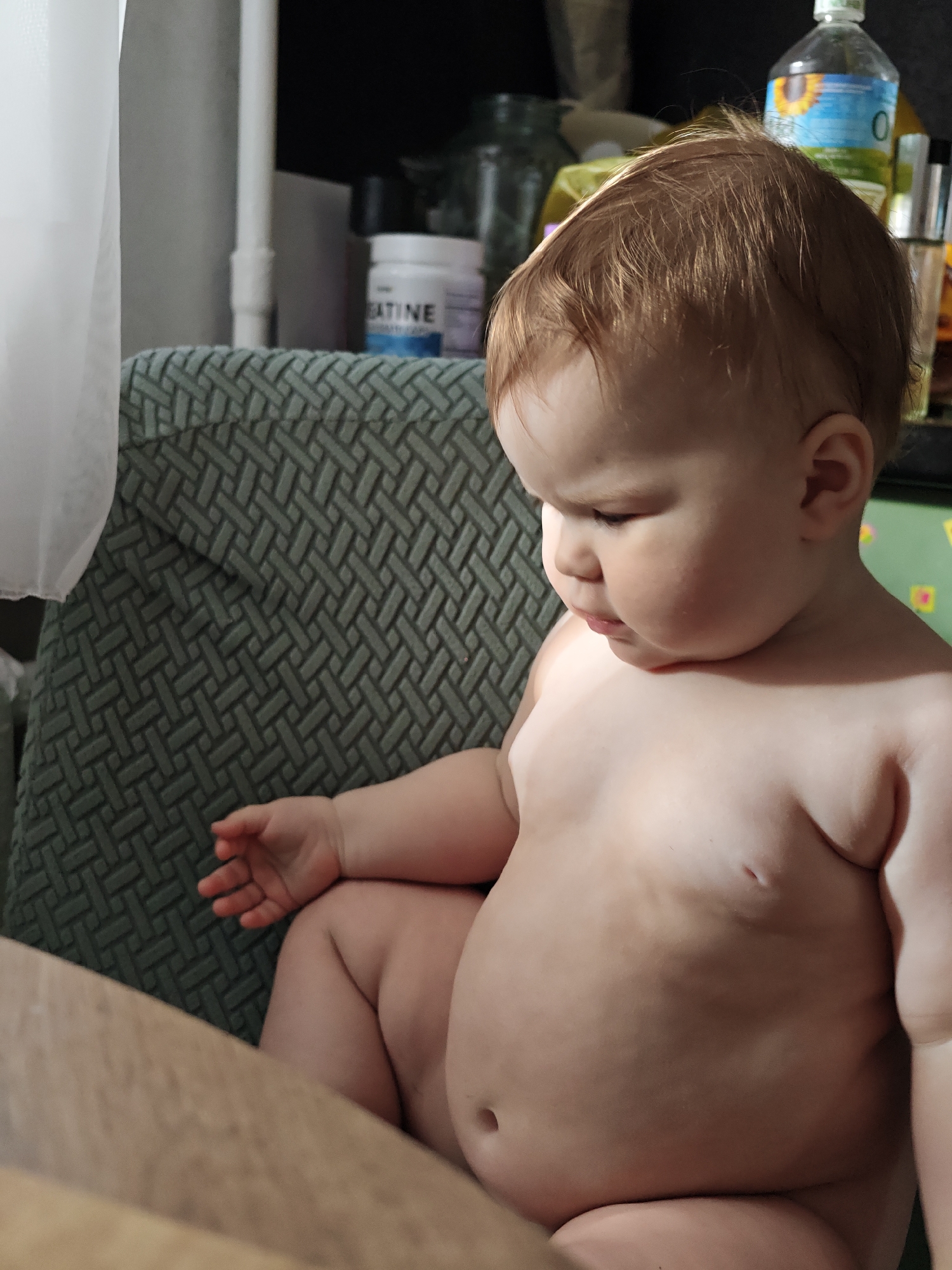 An indoor, close-up shot captures a baby, likely a toddler, with light brown hair, sitting topless or in a diaper. The child is positioned in a chair or high chair featuring a green or teal textured backrest with a prominent geometric, woven pattern. The baby looks down and to their right with a concentrated, contemplative expression.
The background, largely out of focus, reveals several household items. A soft white curtain or fabric hangs on the far left. On a surface behind the baby, a white container prominently labeled "CREATINE" is visible, with further indistinct text beneath it. To the right of the baby's head, a clear bottle with a yellow and blue label featuring a sunflower design can be seen, possibly indicating cooking oil. Other various bottles and jars are also present in the blurred background. In the foreground, a light-colored wooden surface partially obscures the baby's lower body.
The scene depicts a quiet, everyday moment within a home setting in Chaikovsky, Russia, with the baby appearing engrossed in something outside the camera's view. The lighting is soft, suggesting natural light filtered indoors or subtle artificial illumination.