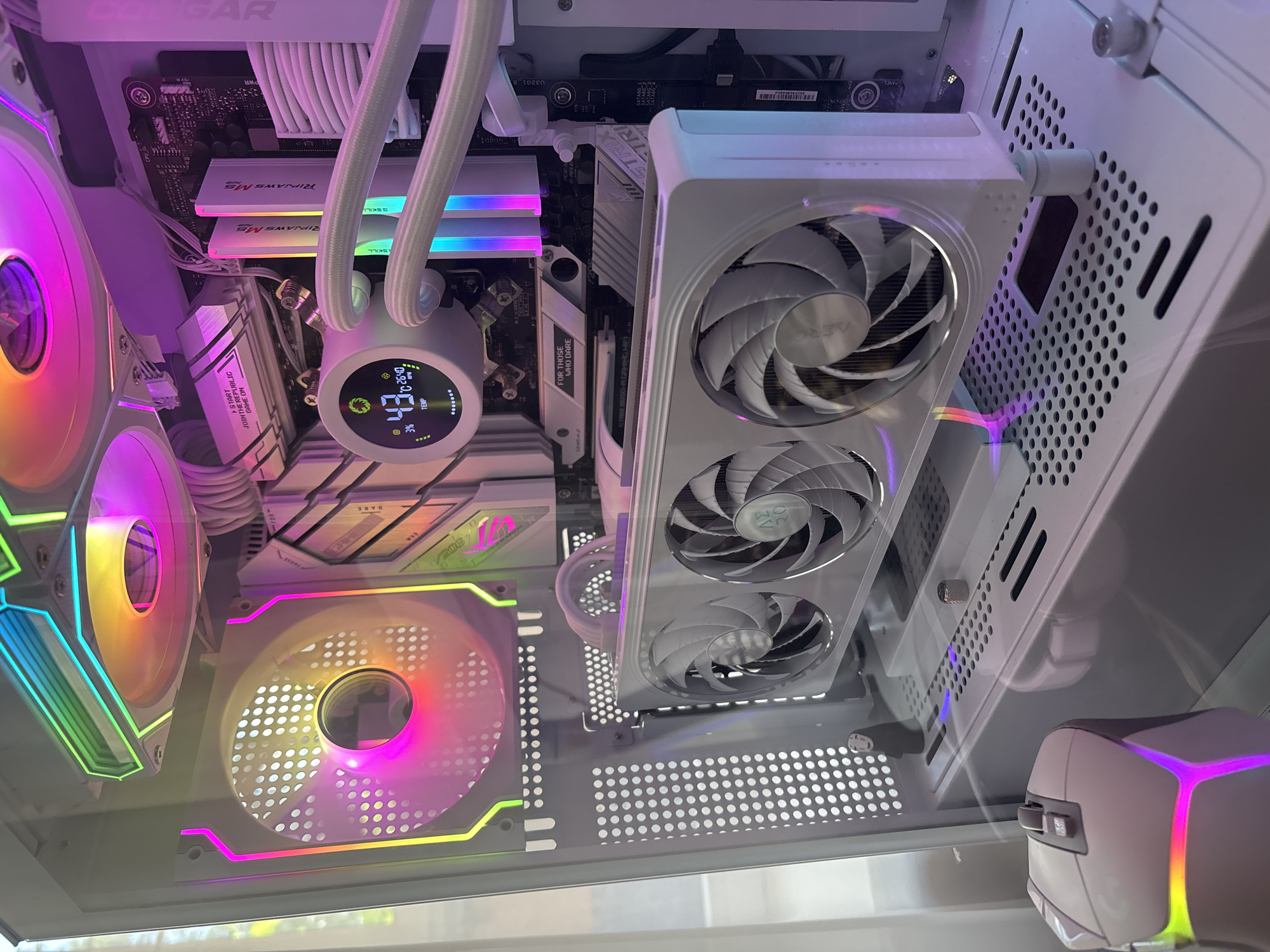 A high-angle close-up shot shows the interior of a white computer tower, illuminated by various RGB lights. The computer features a white GPU with three fans, a white CPU cooler with a circular digital display showing temperature and fan speed, and white RAM sticks. The motherboard and other components are visible, along with white fan cables and tubes. A white gaming mouse with RGB lighting is positioned in the lower left corner of the frame. The background is out of focus, suggesting an indoor environment. The image is taken in an unknown location in Unknown, Unknown.