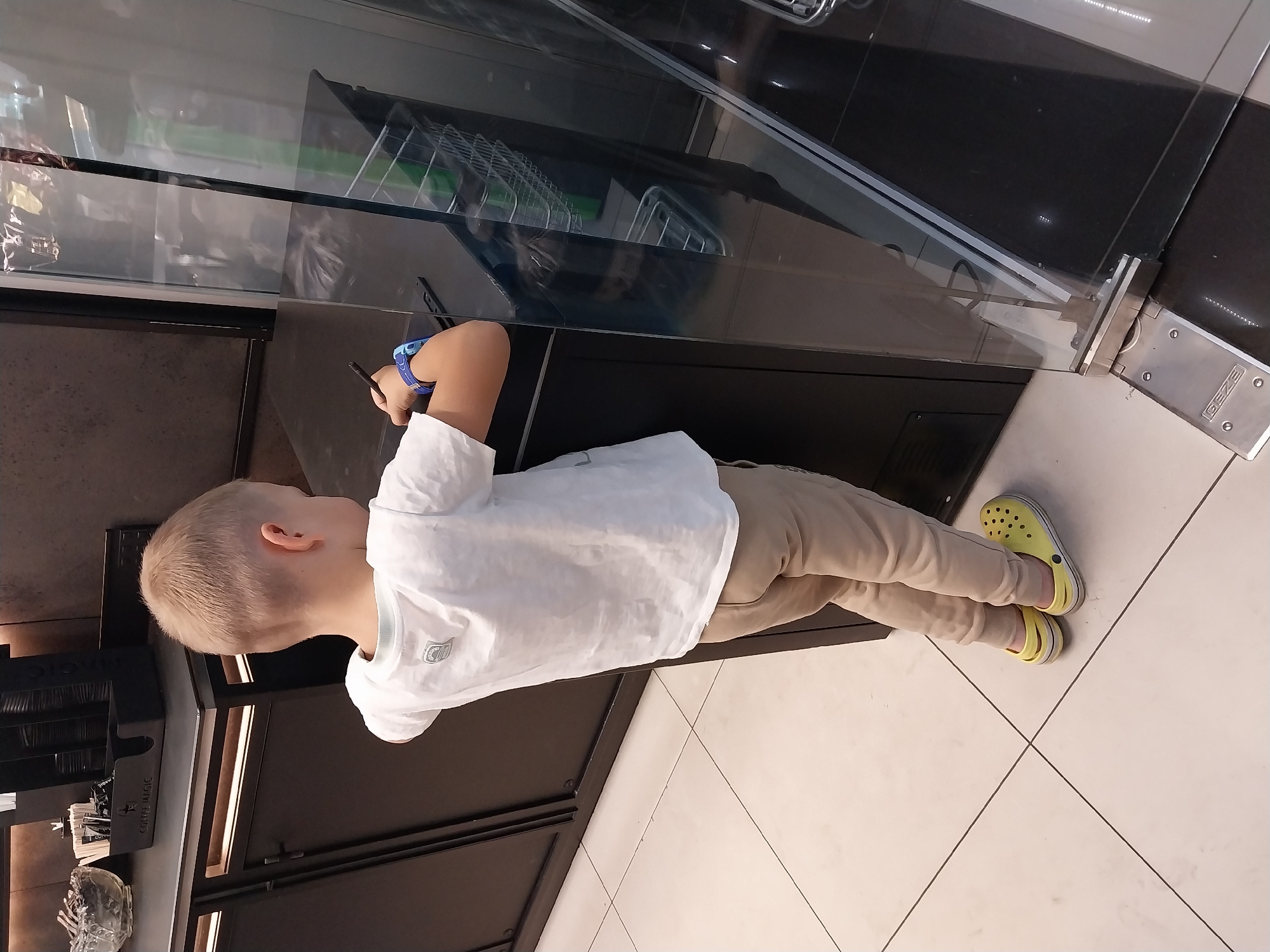 A young boy, viewed from the back, stands in front of a dark counter with a digital drawing tablet and stylus. He is wearing a white t-shirt, tan pants, and yellow clogs. The setting appears to be a public space, possibly a cafe or shop, with tiled flooring and a glass display case to the right. A coffee machine and condiment dispensers are visible on the counter behind the boy. The image was captured in Dnipro, Ukraine.