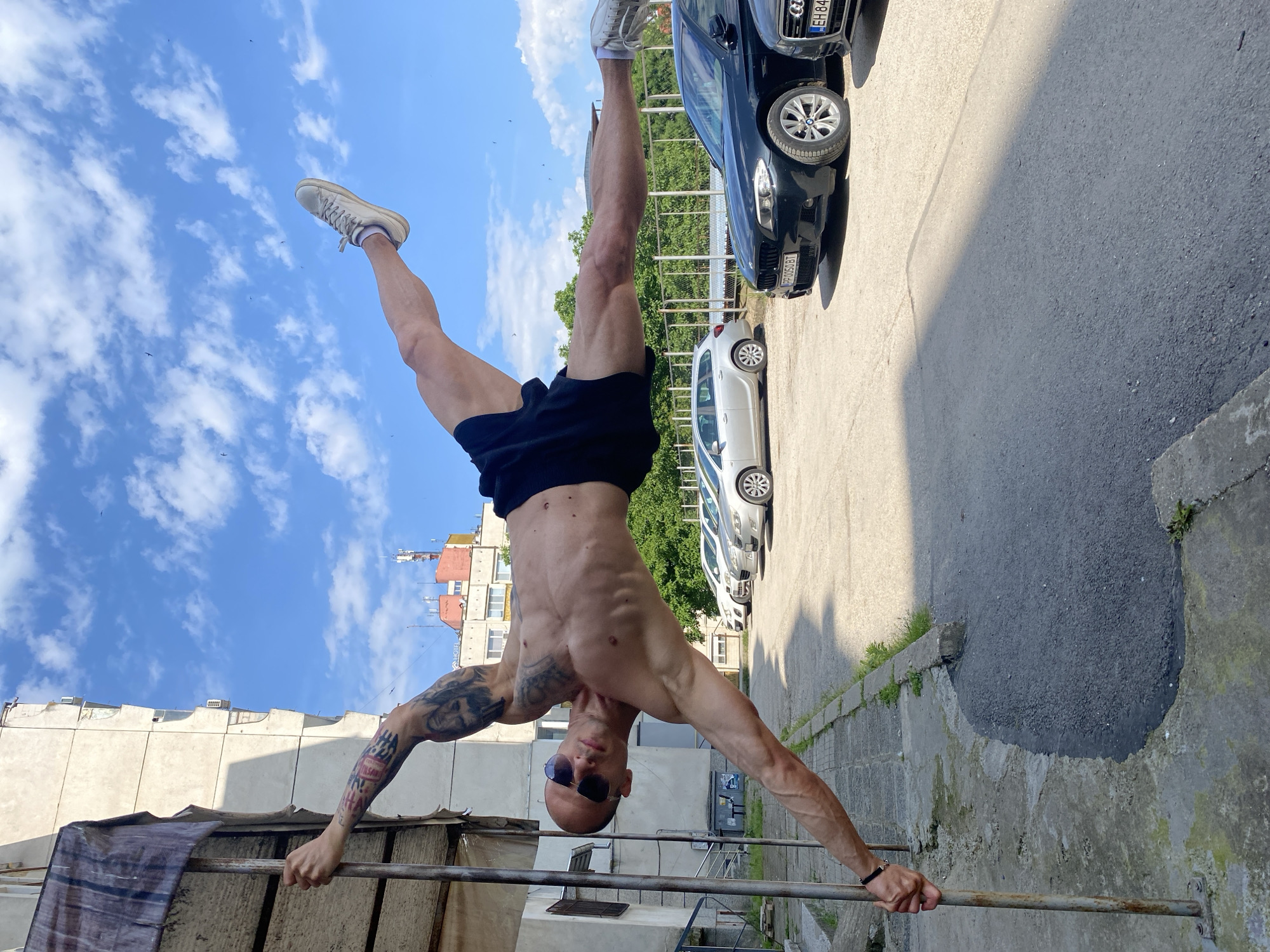 A fit, bald man with tattoos on his arms and a shaved head is performing a human flag exercise, holding onto a metal pole. He is wearing sunglasses and black shorts. His body is horizontal, with one leg extended upwards and the other extended outwards, parallel to the ground. The background shows a residential building, a parking lot with several cars, a chain-link fence, and trees. The sky is blue with white clouds. The image was taken in the city of Pleven, Bulgaria. The license plates visible on the cars suggest the location is Bulgaria.