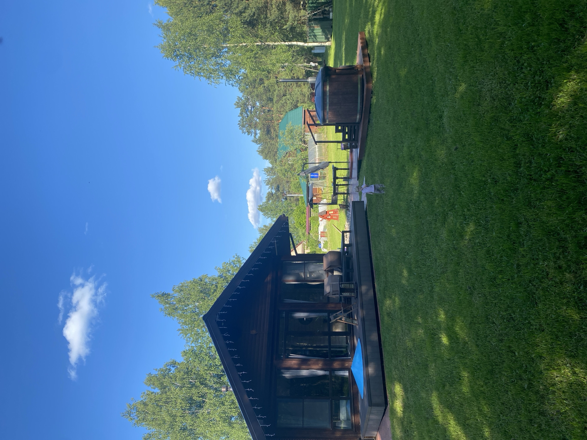 This image presents a serene outdoor scene of a property in Копнино, Russia, under a clear blue sky. The central feature is a charming two-story wooden cabin, characterized by a dark A-frame roof with decorative string lights along its eaves. The cabin boasts large glass windows and doors that reflect the sunny surroundings.
Extending from the front of the cabin is a dark wooden deck furnished with two outdoor chairs. A bright blue mat or towel is placed on the deck floor. To the right of the cabin, on a separate wooden platform, stands a large, barrel-shaped wooden hot tub equipped with an external stovepipe. A small white decorative figurine is positioned on the vibrant green lawn near the hot tub area.
The property is enveloped by a lush, well-maintained green lawn. Various types of trees, including tall pines and lighter green deciduous trees, frame the background, providing a natural and private setting. In the mid-ground, behind the hot tub, a greenhouse or polytunnel with a translucent roof is visible, along with other distant structures.
The scene is bathed in bright sunlight, indicating a clear and pleasant day, likely mid-morning or afternoon. There are no people visible, suggesting a tranquil and unoccupied moment. The overall impression is one of peace and potential for relaxation. No text is visible in the media file.