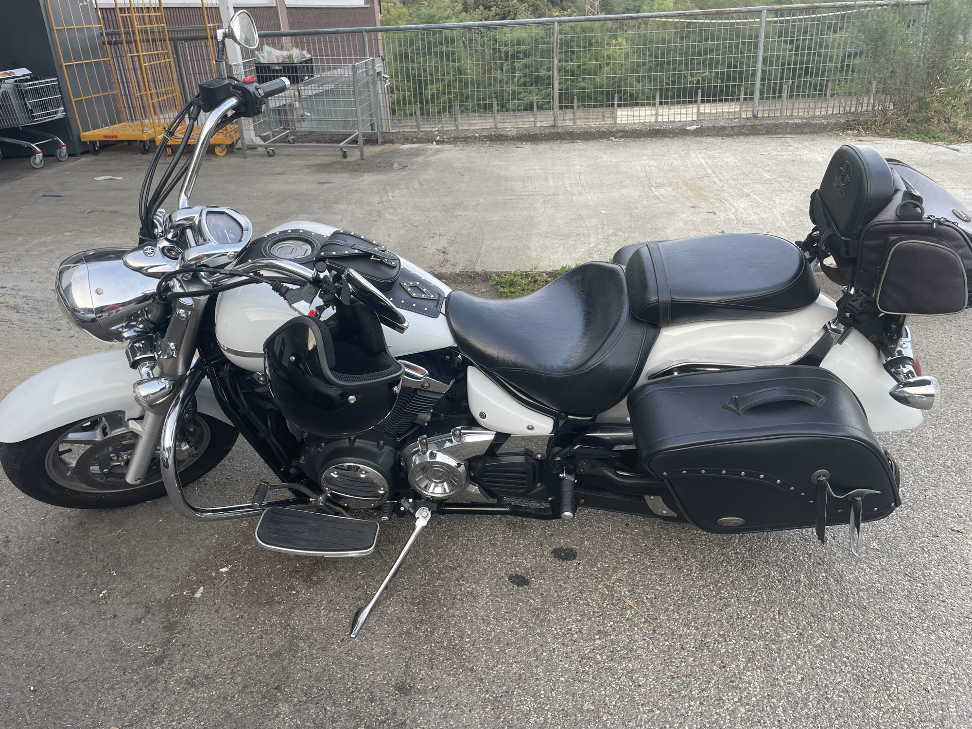 A white and black cruiser motorcycle is parked on an asphalt surface. The motorcycle has a large, black leather seat, black saddlebags, and chrome accents. A black helmet rests on the motorcycle's gas tank. In the background, a metal fence runs along a road, with trees and a building visible beyond the fence. Several shopping carts are also present in the background. The scene appears to be an outdoor area, possibly a parking lot. The location is identified as Borgosesia, Italy.