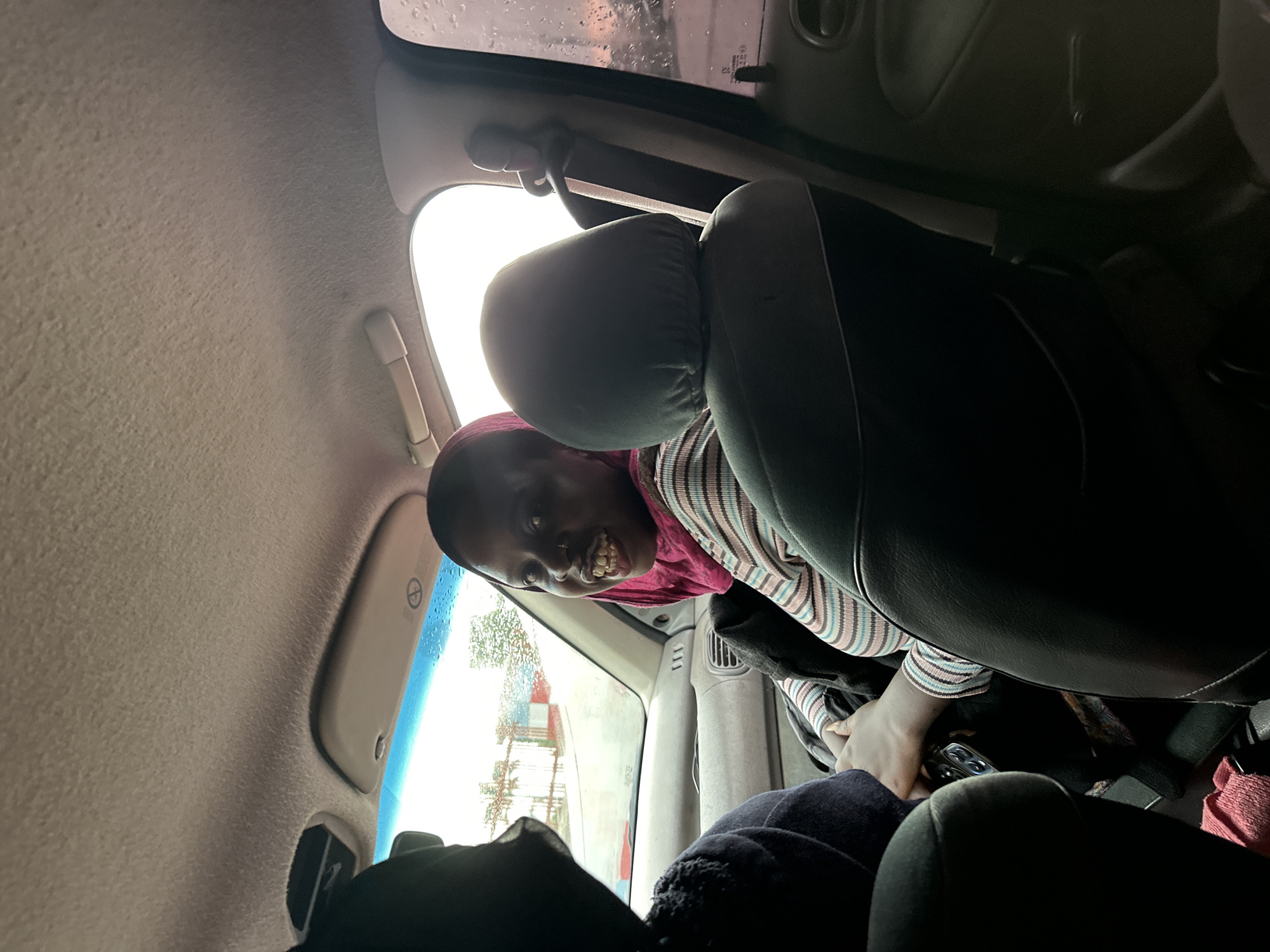 The media file shows a young person looking directly at the camera from inside a car. They are wearing a pink headscarf and a striped long-sleeved shirt. Their mouth is open in a wide smile, showing their teeth, and their eyes are wide. They appear to be in the back seat of the car, leaning forward towards the camera.
The interior of the car is visible, including the car's headrests and ceiling. The window shows a cloudy sky with raindrops on it, suggesting it may be raining or has recently rained. Outside the car, through the window, there are glimpses of buildings and trees. The overall scene conveys a sense of lightheartedness and engagement with the person taking the photo. The setting appears to be an urban or semi-urban environment in Gura Top, Nigeria. There are no visible text elements in the image.
