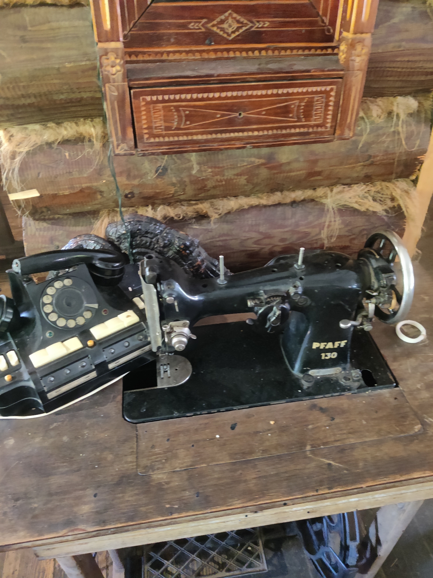 The image shows a close-up, eye-level view of a vintage Pfaff 130 sewing machine positioned on a wooden table. To the left of the sewing machine is a black rotary dial telephone, also appearing to be of vintage design. Both objects are situated in a rustic indoor setting, with exposed wooden beams and what appears to be straw or similar material visible in the background, suggesting a rural or historical location. Above the objects, a decorative wooden structure with intricate carvings is mounted on the wall. The lighting is moderate, highlighting the textures of the wood and the machinery. The scene is static and appears to be a still life arrangement of these items. There are no people or animals present in the image. The location is identified as Izky, Ukraine.