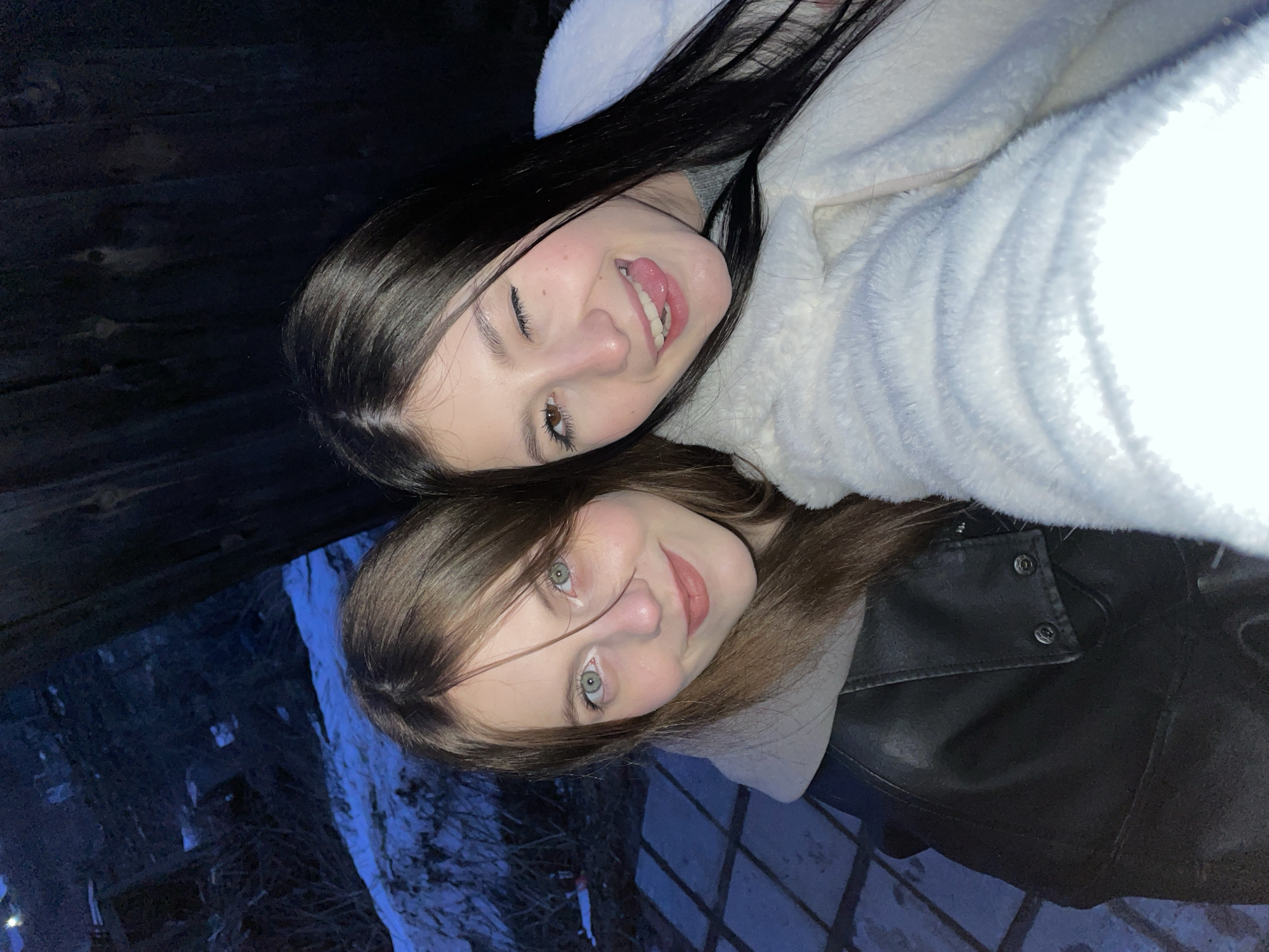 Two young women are posing for a selfie outdoors. One has long, dark hair and is wearing a white, furry jacket; she is sticking her tongue out and winking. The other has shoulder-length brown hair and is wearing a black leather jacket over a white hoodie. The background appears to be a dark, wooded area, possibly with snow on the ground, near Фоки, Russia. A section of the background shows what appears to be a man-made wooden structure.