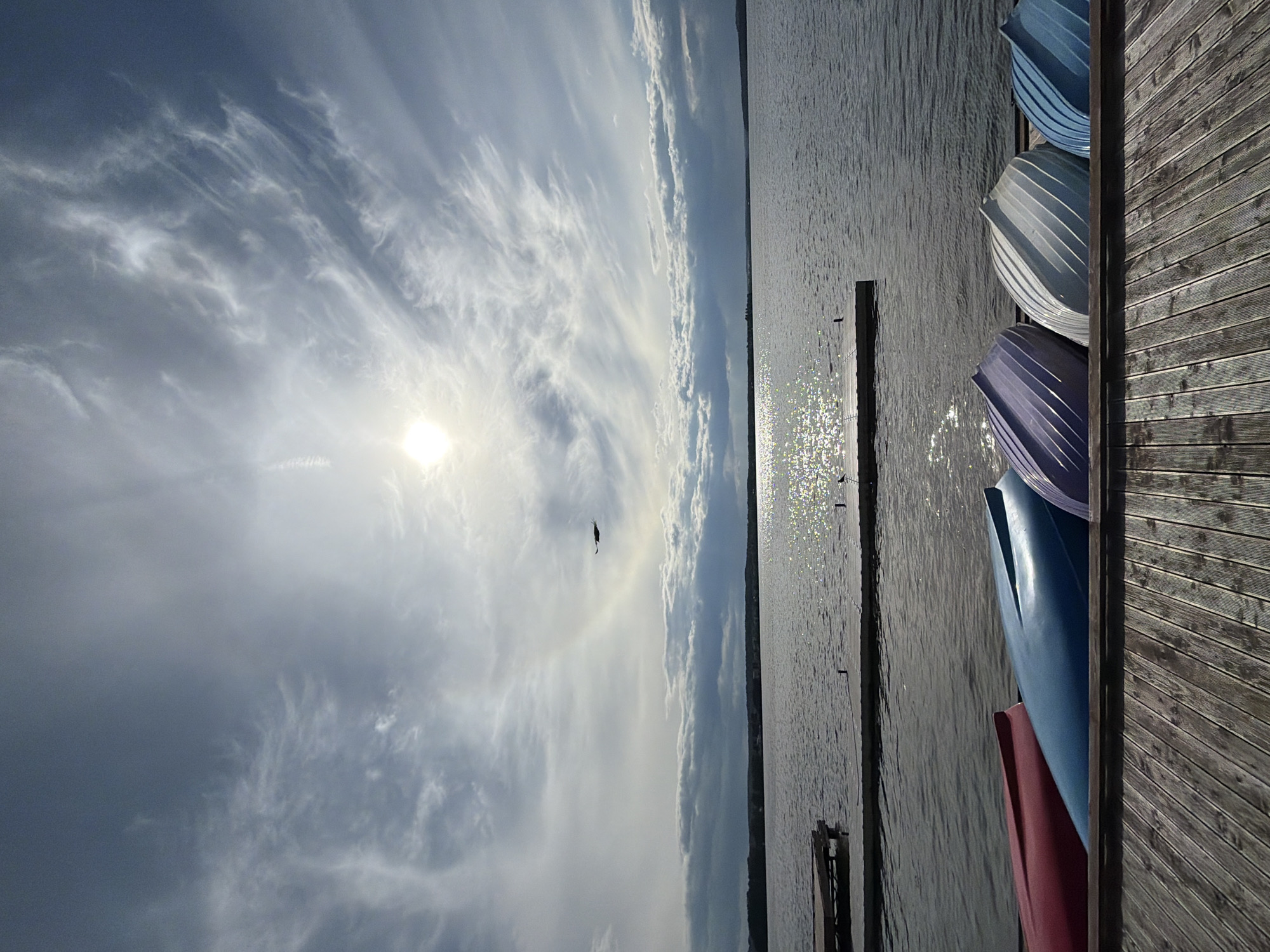 A view of a lake in Votkinsk, Russia, shows several colorful boats resting on a wooden dock in the foreground. The sky above is partly cloudy with the sun visible, casting a reflection on the water. A bird is in flight.