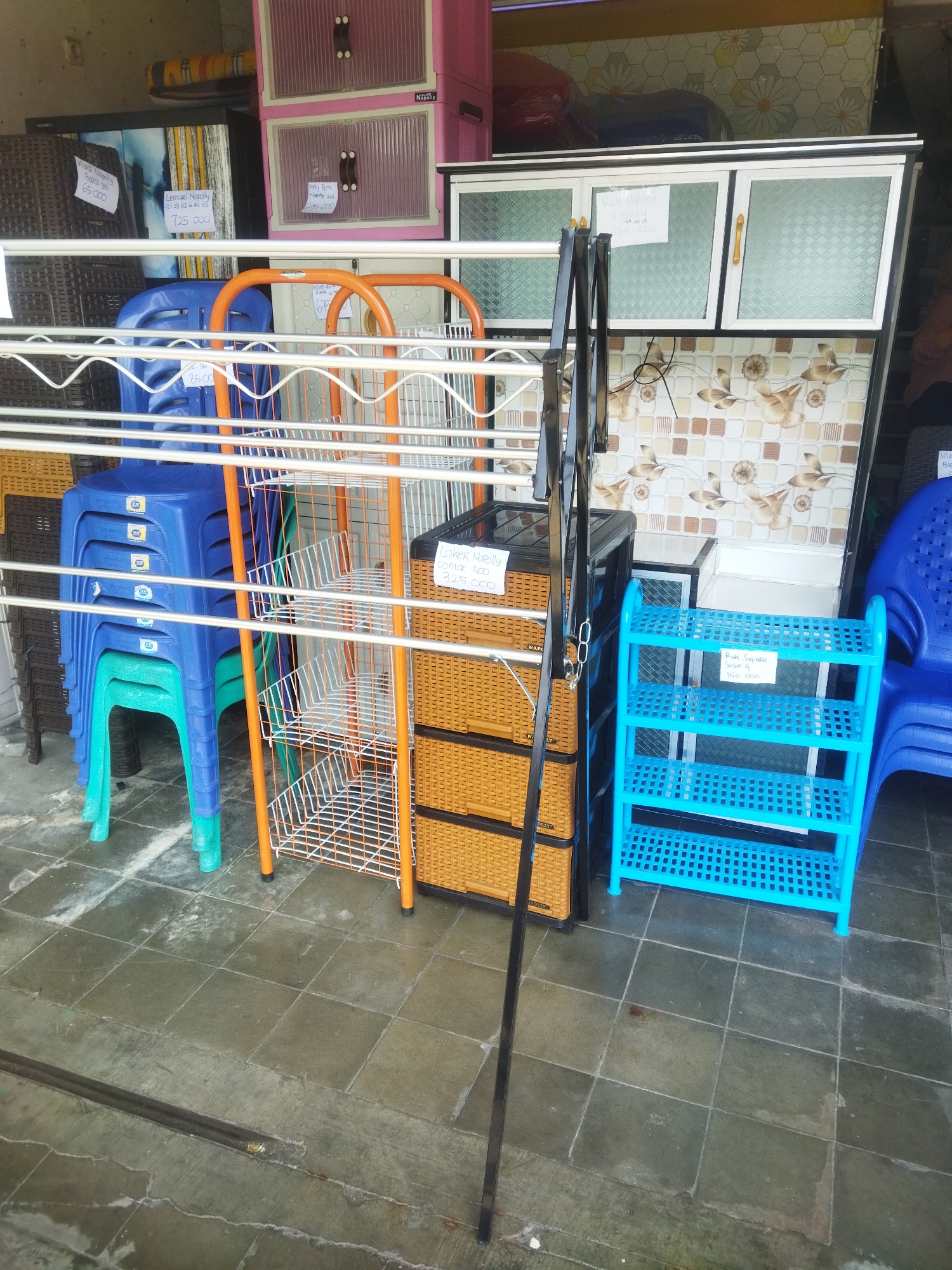 This is a medium shot, eye-level, outdoor, daytime, full-color photograph of a retail storefront in Wonogiri, Indonesia, displaying various plastic storage and seating items. The background features a light-colored tiled wall with a floral pattern, a white cabinet with glass doors, and a pink and white stacked plastic cabinet. In the foreground, several items are arranged on the tiled floor. To the left, a stack of blue plastic chairs with teal legs are neatly piled. Next to the chairs is an orange metal shelf unit with multiple white wire baskets. Adjacent to the orange shelf is a brown woven plastic chest of drawers. To the right of the chest of drawers is a bright blue plastic shelving unit with four tiers. A metal drying rack with black legs stands in the center, partially obscuring the items behind it. In the upper right corner, a blue plastic chair is visible. Several price tags are attached to the displayed items, some of which are legible. The overall scene suggests a storage and furniture shop.