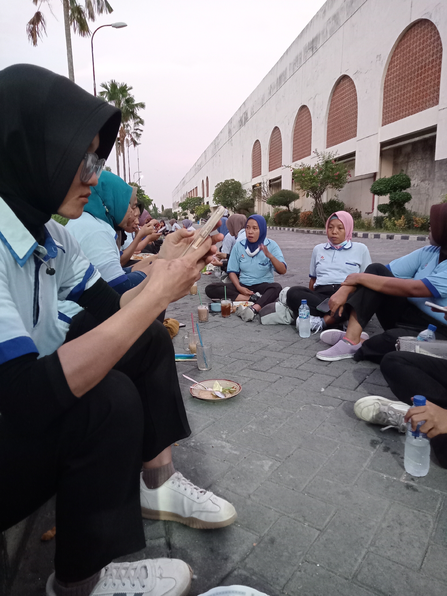 A group of approximately seven women are seated on a paved surface, possibly a parking lot or walkway, in what appears to be an outdoor setting. The women are wearing light blue shirts with dark blue collars and long black pants. Many are wearing hijabs of various colors. Several women are using cell phones, while others are eating or drinking. Empty plates, cups, and water bottles are scattered around them on the pavement. In the background, a large white building with arched windows and several trees and shrubbery are visible. Palm trees and street lights are also present in the background, under a dusk or overcast sky. Based on the evidence that the location is Sukomoro, Indonesia, the scene depicts a casual social event or a meal break outdoors for these women.