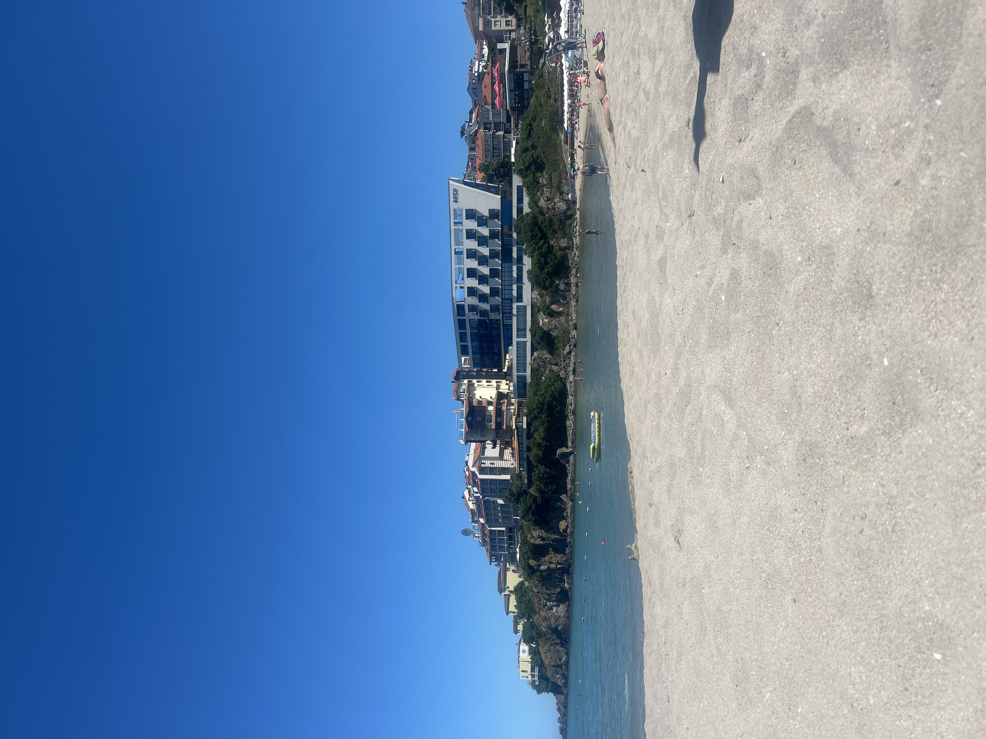 A wide-angle shot of the beach in Sozopol, Bulgaria, shows a clear blue sky above a vibrant coastal town. The sandy beach in the foreground is mostly empty, with a few people sunbathing and walking near the water's edge. The water is calm and blue, with a few buoys marking an area of the sea. In the distance, a cluster of buildings, including hotels and residential properties, are built on a rocky incline overlooking the sea. The architecture of the buildings ranges from modern, with large windows and balconies, to more traditional, with red-tiled roofs. Some of the buildings feature the word "SOZOPOL" on their facade. The overall scene depicts a serene summer day at a popular tourist destination.