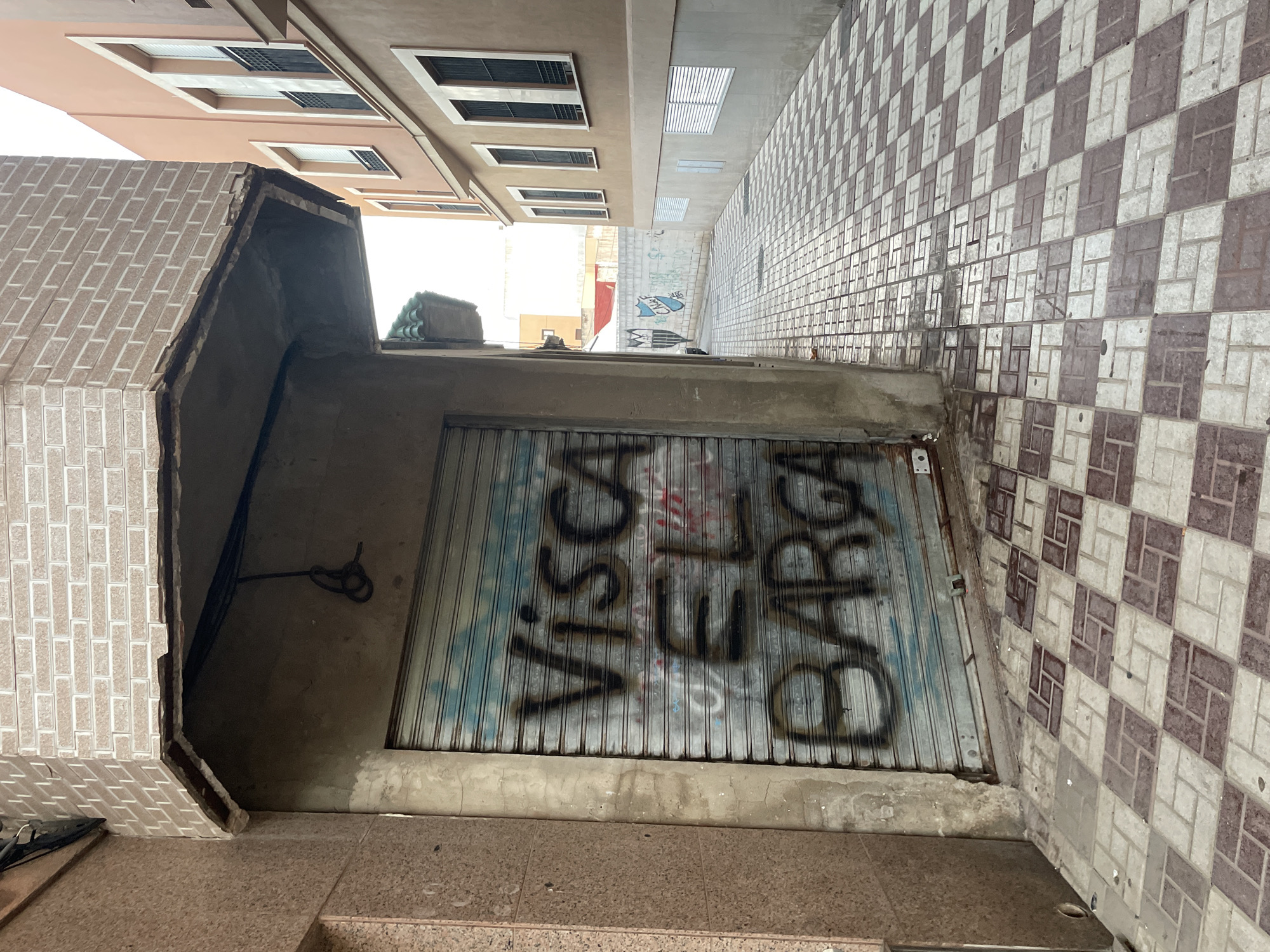 The image depicts a narrow street in La Línea De La Concepción, Spain, with a prominent, closed metal shutter on the left, covered in graffiti. The graffiti appears to say "Visca El Barca". The shutter is part of a building with textured, reddish-brown facade panels to its left and a concrete pillar. Above the shutter, a damaged concrete overhang juts out, showing exposed interior materials and electrical conduits. The ground is paved with a checkerboard pattern of light and dark red square tiles. The street opens up to the right, revealing more buildings. The building on the right has a beige facade with multiple windows, each featuring black bars and white shutters or frames. The street continues into the distance, where more buildings are visible, and a white wall with blue graffiti, including a shield-like emblem, can be seen. The sky is overcast, and the lighting suggests daytime. There are no people visible in the image.