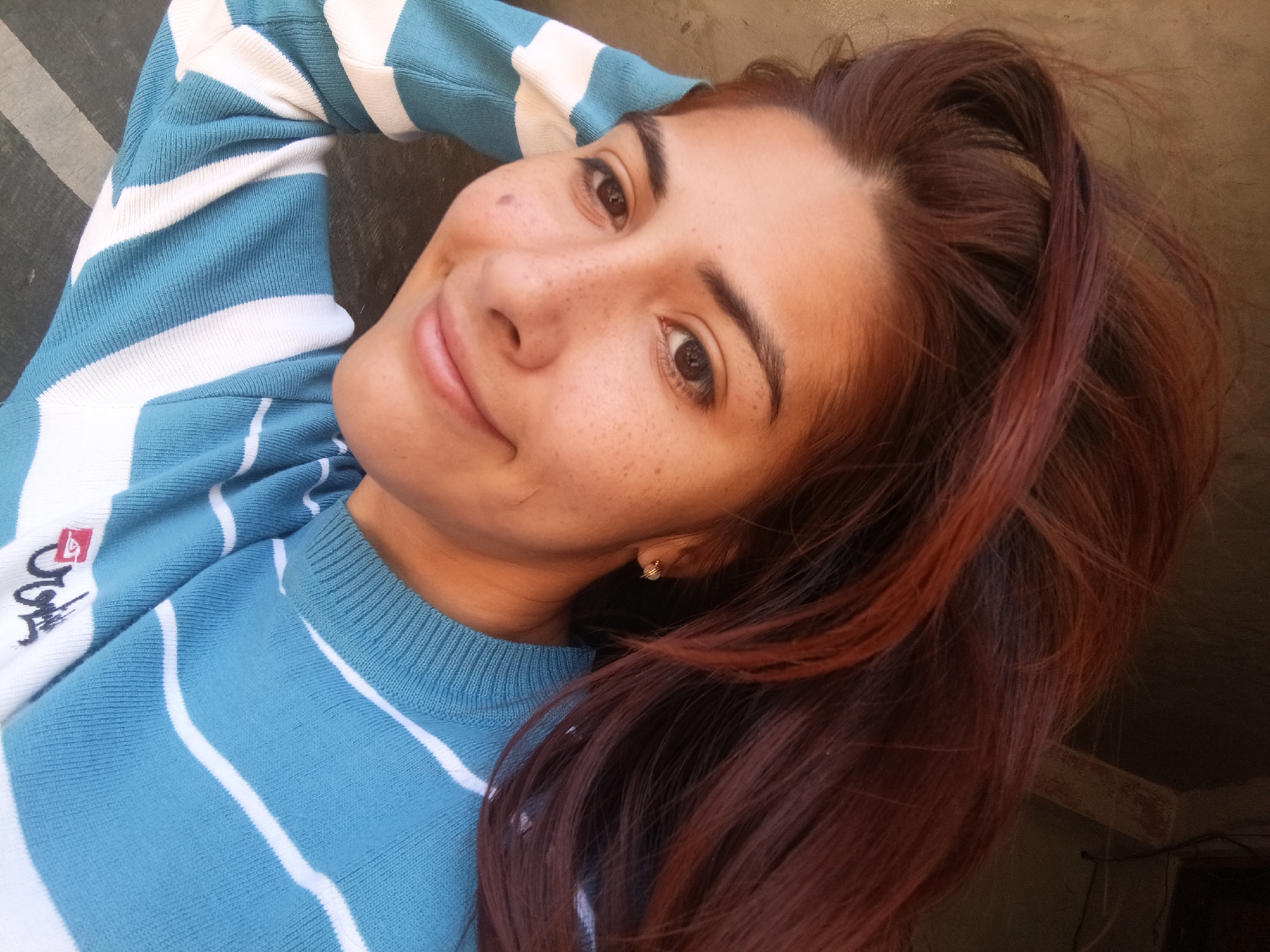 A young woman with auburn hair poses for a selfie. She is wearing a light blue and white striped sweater. She has light skin with freckles and brown eyes. Her hair is partially pulled back. A small gold earring is visible in her left ear. The background is blurry, with the corner of a wall visible in the lower right. The location and its environment is undetermined.