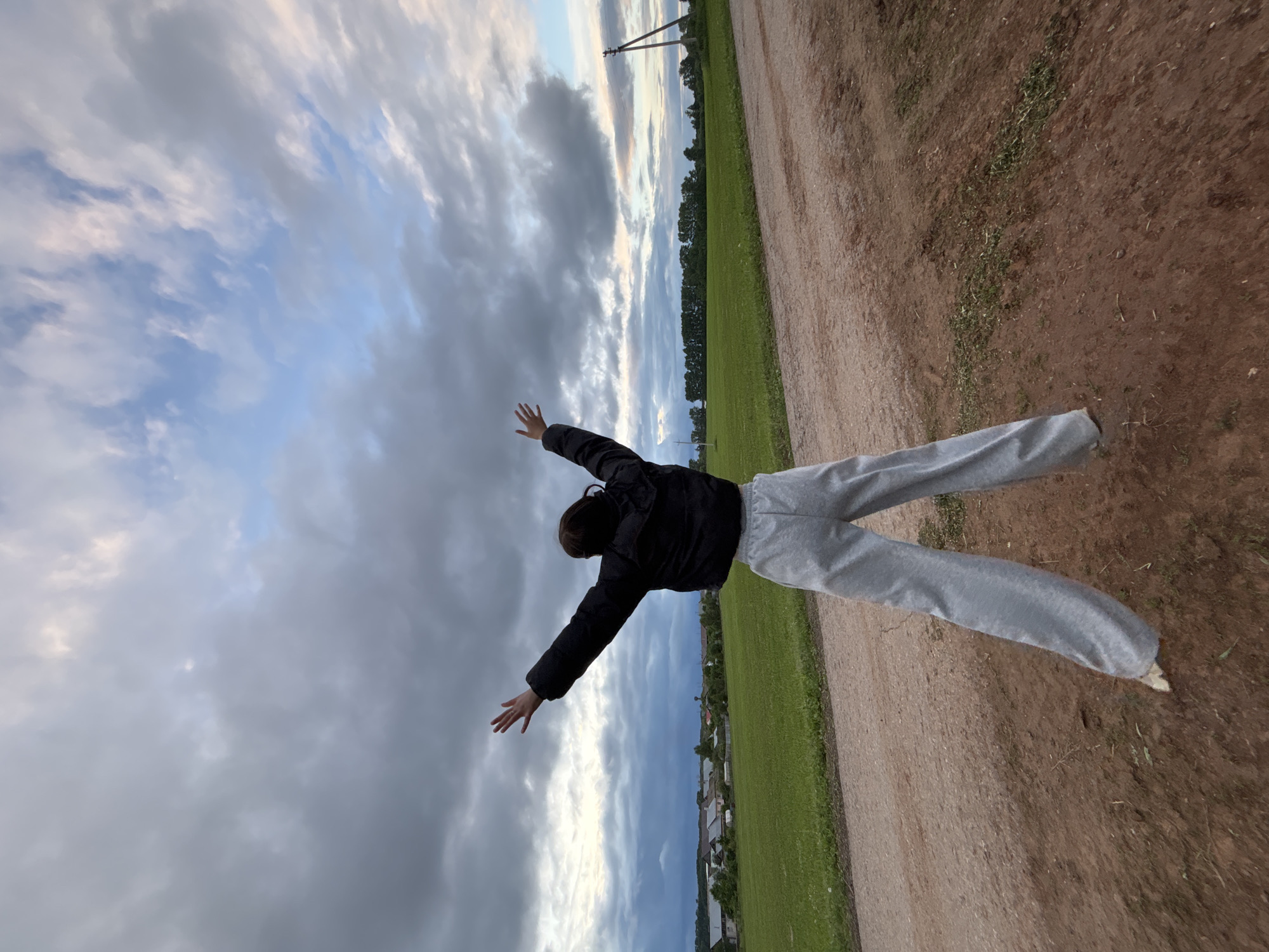 The image depicts a rural landscape in Фоки, Russia. A single individual, viewed from the back, stands on a reddish-brown dirt path. The person is wearing a black cropped puffer jacket and light grey wide-leg sweatpants, with white footwear partially visible. Their arms are raised and spread wide, with palms open and facing upwards, as if embracing the environment. The dirt path in the foreground exhibits some unevenness and scattered green vegetation. On either side of the path, expansive green fields stretch towards the distant horizon. To the mid-left, a small settlement or village with several buildings featuring light-colored roofs is visible among trees. Further right, a dense line of trees forms a dark silhouette against the sky, and two wooden utility poles, one significantly leaning, are visible on the far right. The sky above is largely overcast with a dynamic arrangement of dark grey, lighter grey, and hints of pale blue and warm white clouds, suggesting a transitional period of day such as dawn or dusk.