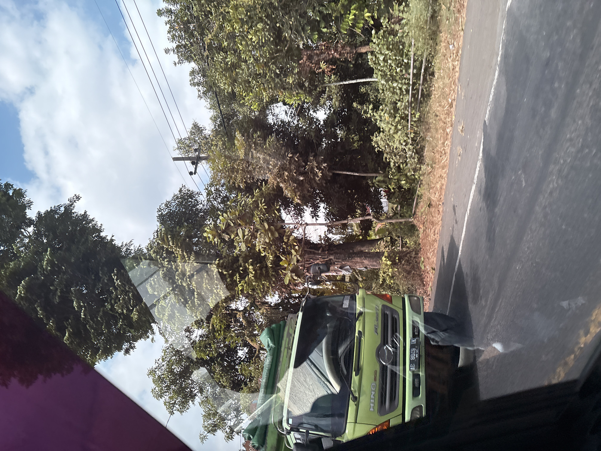 The image shows a green Hino truck driving on a road in Gambirono, Indonesia. The truck is loaded with green tarps. The road is paved and has a white line down the center. Trees and vegetation line the side of the road. The sky is blue with some white clouds. The image appears to be taken from inside another vehicle, as evidenced by the reflection on the glass.