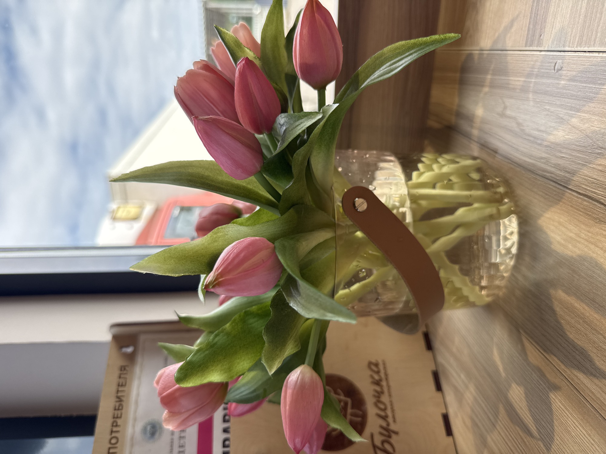 A bouquet of pink tulips is displayed in a clear glass vase on a wooden surface. The vase has a tan leather handle attached to it. Behind the tulips, a cardboard box with Cyrillic text is visible. The background shows a window with a view of a cloudy sky and buildings. The overall scene is brightly lit, suggesting it is daytime. The image captures a still life arrangement in what appears to be an indoor setting, possibly a cafe or shop, given the context of the "Finskiy Briz" location in Russia.