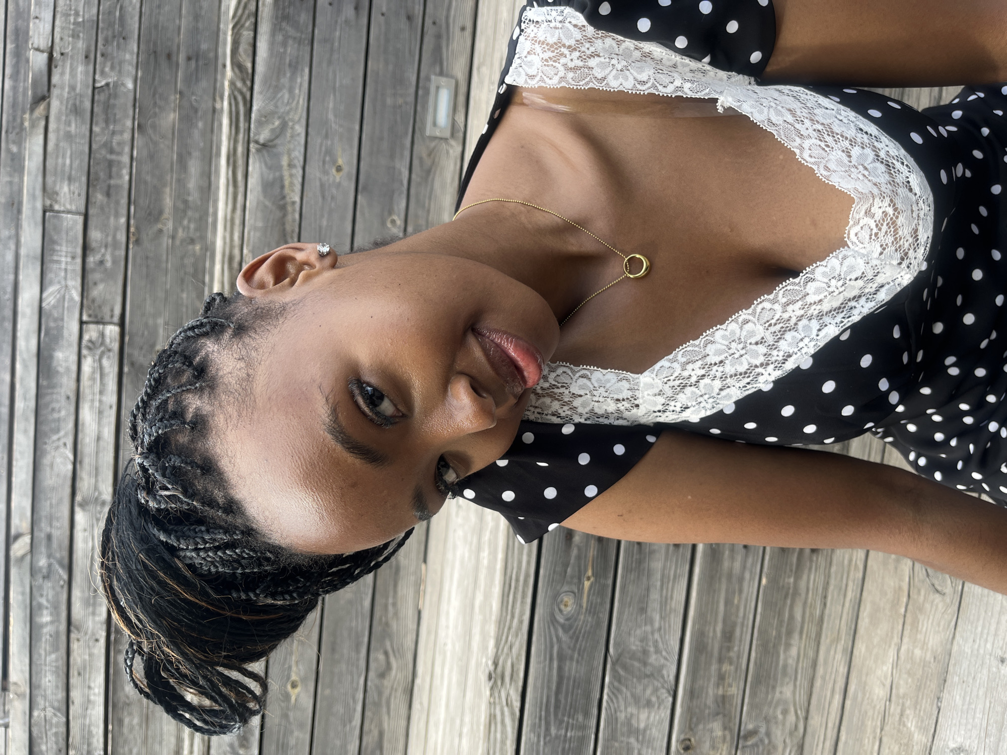 A young Black woman with dark braided hair is captured in a close-up portrait. She is wearing a black and white polka dot dress with white lace trim along the neckline. A thin gold necklace with a circular pendant rests on her collarbone. She has a stud earring in her visible ear. Her head is tilted, and she is looking towards the camera with a slight smile. The background consists of weathered wooden planks, likely an outdoor setting.