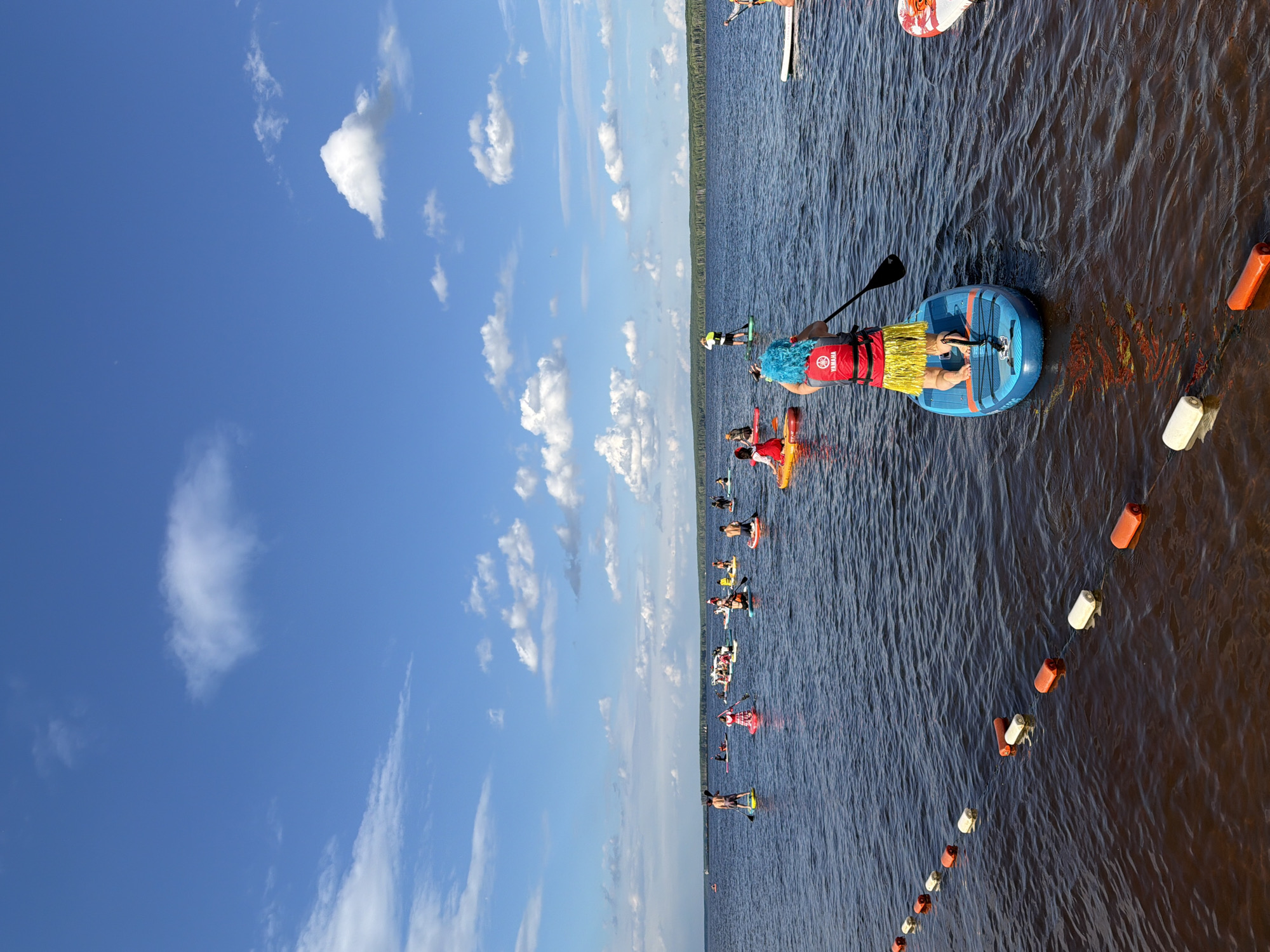 The image depicts a group of people paddleboarding on a body of water. The scene is set on a sunny day with a bright blue sky filled with scattered white clouds. The water appears calm with gentle ripples. A line of orange and white buoys is visible in the foreground, marking a course or boundary on the water.
The paddleboarders are positioned at various points on the water, with some closer to the viewer and others further in the distance. Many are wearing life vests, and some have on colorful wigs and costumes, suggesting a festive or recreational event. One person in the foreground is wearing a red life vest and a yellow grass skirt, with a bright blue wig. Another paddleboarder behind them is wearing a similar costume.
The activity appears to be a paddleboarding race or a group paddleboarding event. The paddleboarders are all facing in the same general direction, indicating they are moving together. The shoreline, lined with trees, is visible in the background, suggesting a natural setting. The overall mood seems lighthearted and fun, with the costumes adding a playful element to the event. The presence of buoys implies a designated area for the activity.
There is visible text on one of the paddleboards: "YAMAHA". There is also text on a red and white paddleboard in the upper right portion of the image, which appears to read "AVANTI". The time of day seems to be daytime, likely late morning or afternoon, given the bright sunlight and clear skies. The location is identified as Chaikovsky, Russia.