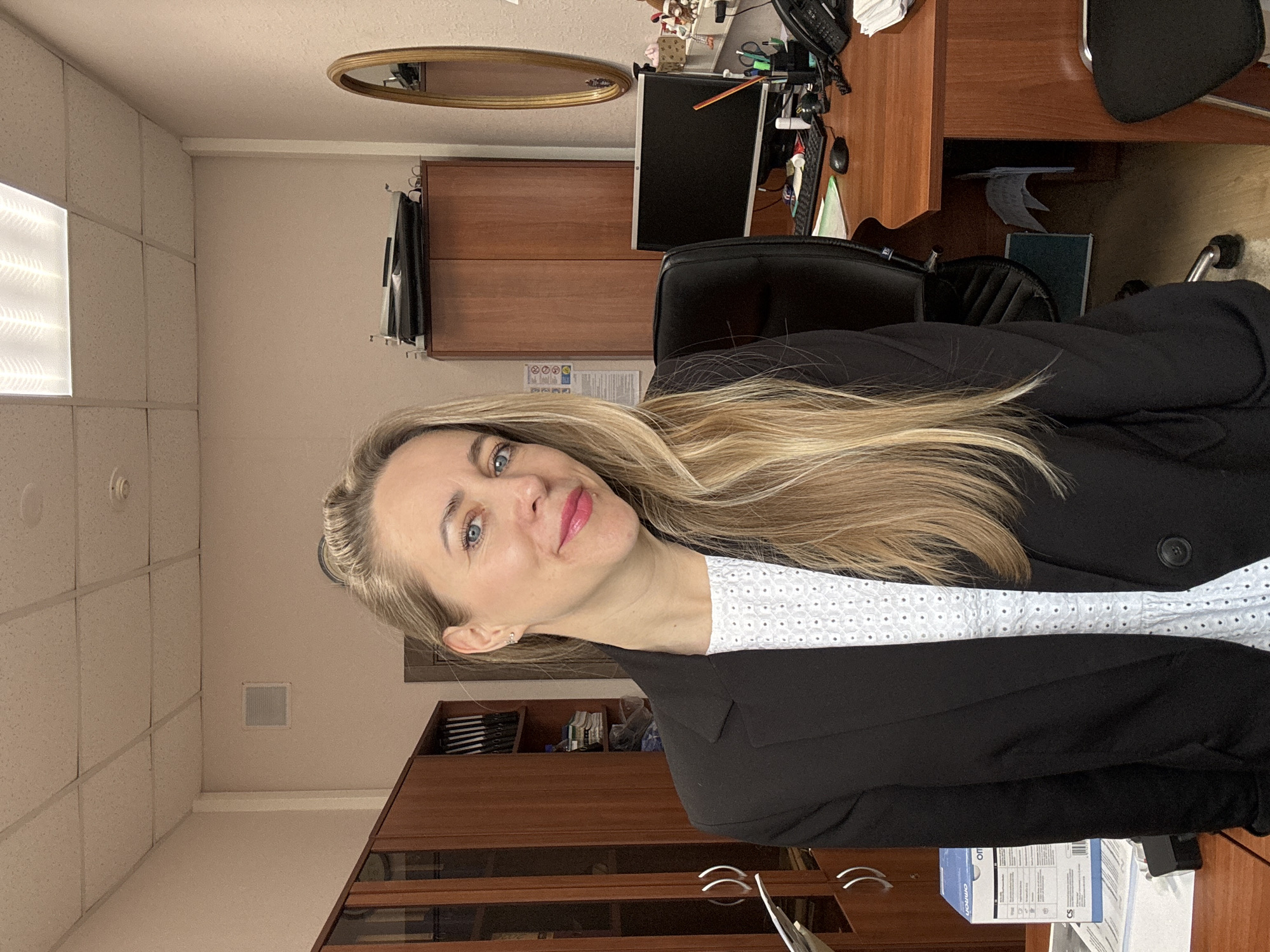 The image is a medium shot of a woman with blonde hair, wearing a black blazer over a white patterned shirt. She is looking at the camera with a slight smile.
Behind her is an office setting, with a wooden desk and chair. On the desk, there is a computer monitor, keyboard, and various office supplies. A wooden cabinet is visible behind her. The office is located in a building with a white tile ceiling and beige walls. It is not possible to determine the precise location in the city of Chaikovsky, Russia, from the image alone.