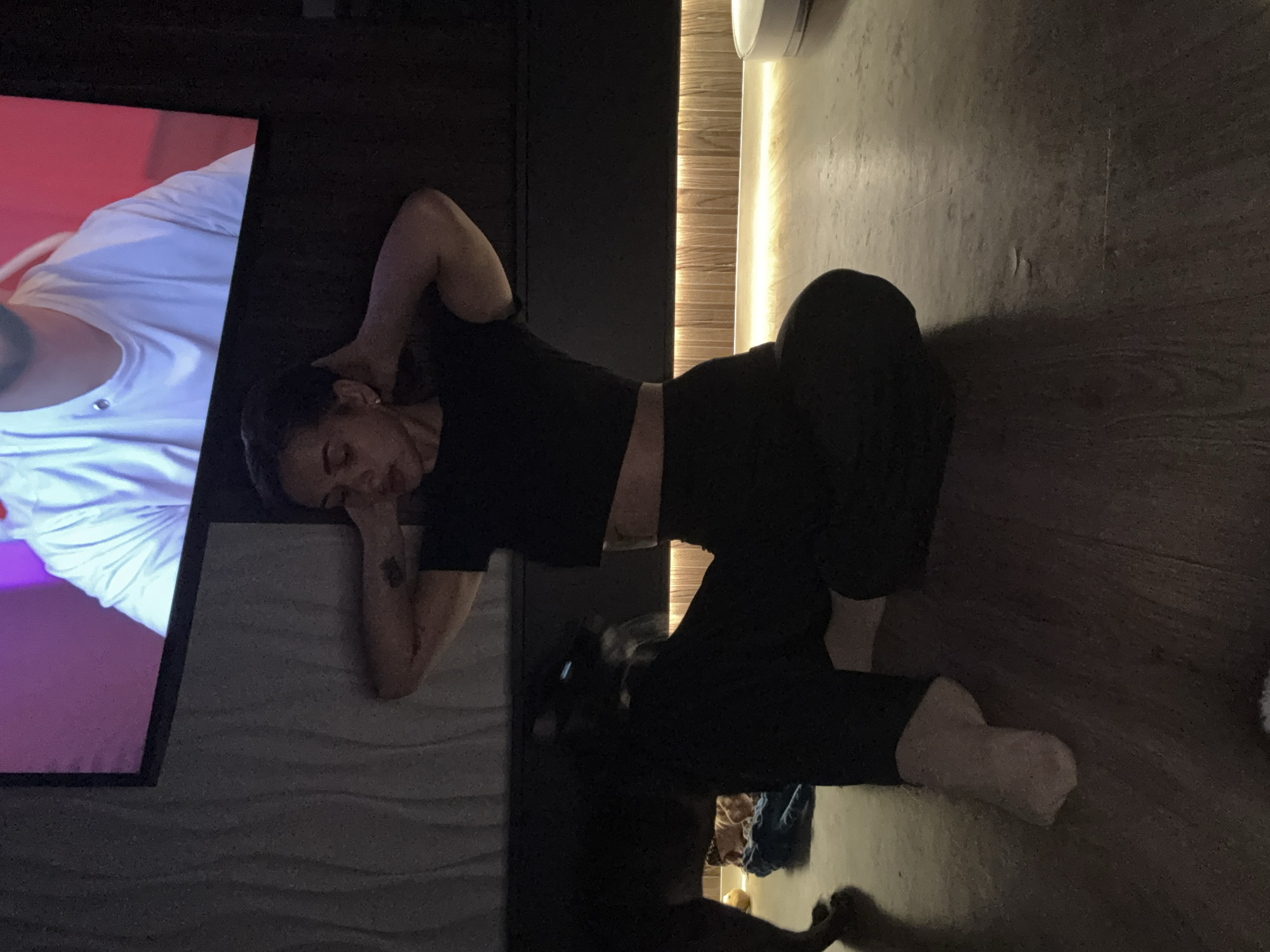 A person lies on their side in a darkened room, wearing a black crop top and black pants. Their head rests on their hand, with an arm bent behind their head. A television screen displays a person in a white shirt with a purple and red background. The person is lying on a textured surface next to the television. A partially visible object resembling a clothing rack or storage unit is behind the person. The location cannot be determined to be in Лобаново, Russia, based on the image alone.