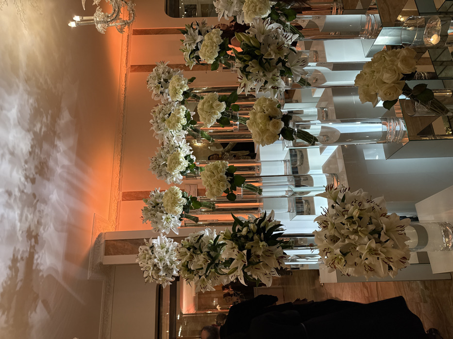 A cascading display of white floral arrangements lines a mirrored staircase. Cylindrical glass vases hold the bouquets, featuring lilies, roses, and other white blossoms. The stairs have a multi-tiered, reflective design. Soft, warm lighting illuminates the scene, creating a glamorous ambiance. Part of a chandelier is visible in the upper left.