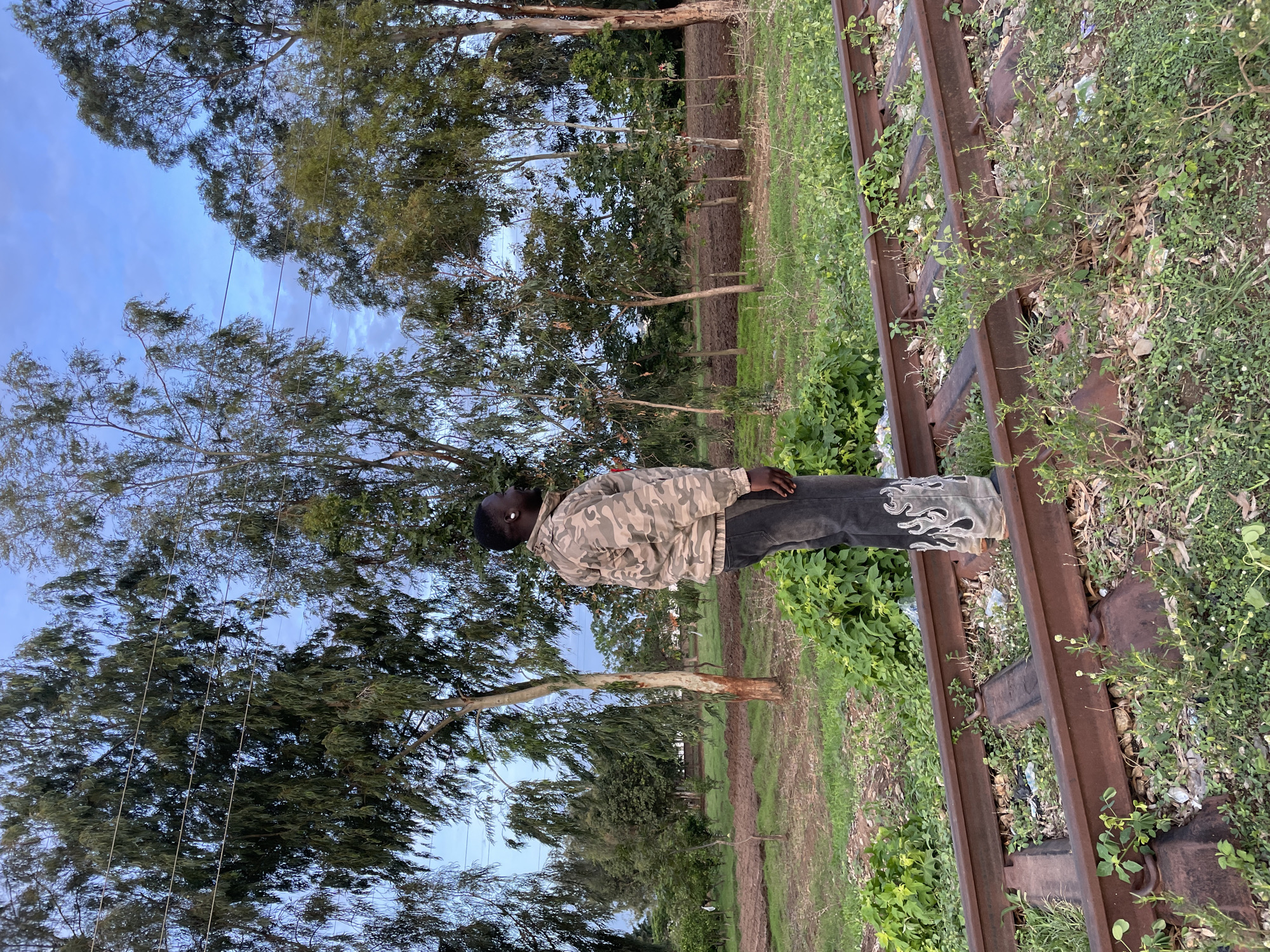 The image depicts an outdoor scene in Gura Top, Nigeria, featuring a lone individual positioned on disused railway tracks amidst a natural environment. One male individual with dark skin is shown in profile, facing towards the right side of the frame. He is wearing a camouflage-patterned jacket in shades of tan, green, and brown, and dark-colored jeans adorned with a white flame-like design on the lower outer legs. A white earbud is visible in his right ear. The individual's right foot appears to be on or directly next to one of the railway rails, while his left foot is on the grassy ground adjacent to the tracks.
The railway tracks, consisting of two rusty metal rails connected by sleepers, traverse the lower portion of the image. Significant vegetation, including small plants, weeds, and grass, grows between and around the rails, indicating infrequent use or disuse. Small pieces of litter are visible on the ground around the tracks.
The environment is characterized by abundant greenery. Numerous trees of varying heights and foliage types dominate the midground and background, forming a dense canopy against the sky. Some trees feature long, drooping leaves. The ground is a mix of green vegetation and reddish-brown soil, with patches of tilled earth visible further in the background, possibly indicating agricultural activity. Thin, dark lines, resembling power or communication wires, are stretched across the sky between the trees. The sky is partially cloudy with visible patches of light blue, suggesting either early morning or late afternoon lighting conditions.
