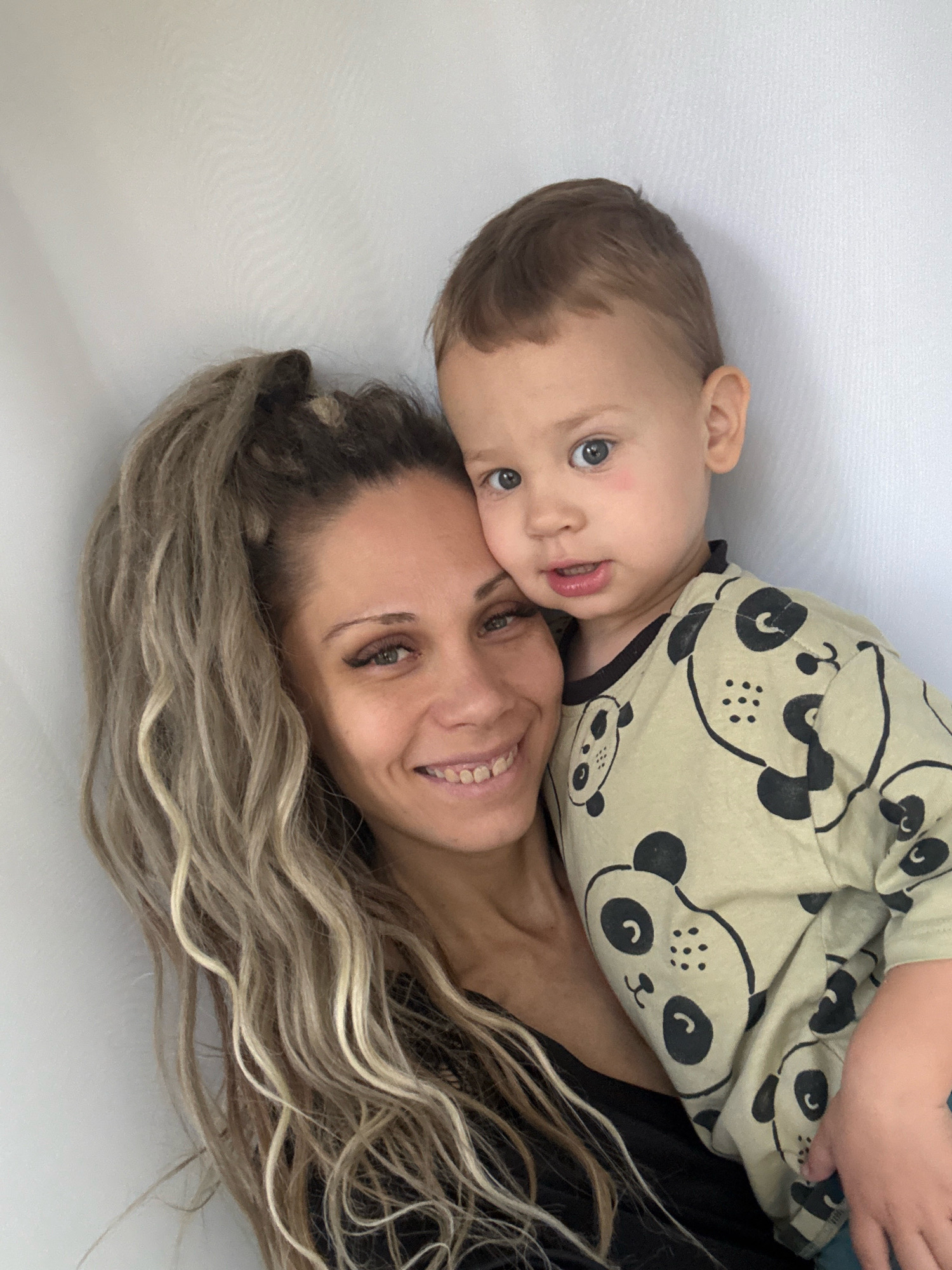 A medium close-up shot captures a mother and her young child against a plain, textured white background, possibly a wall. The mother, positioned on the left side of the frame, is looking towards the camera with a warm smile and her eyes are open and bright green. Her long, blonde, wavy hair is pulled up into a high ponytail, with several strands falling loosely around her face. She is wearing a black top. The child, a toddler with fair skin and light brown hair, is nestled in the mother's arms and looks directly at the camera with wide, blue eyes. The child is wearing a light green t-shirt adorned with a repeating pattern of black panda faces. The mother's left arm is visible, holding the child. The overall lighting is soft, and the image appears to be taken indoors. There is no indication of the city of Rusanovo, Russia, or any specific urban environment in the image.