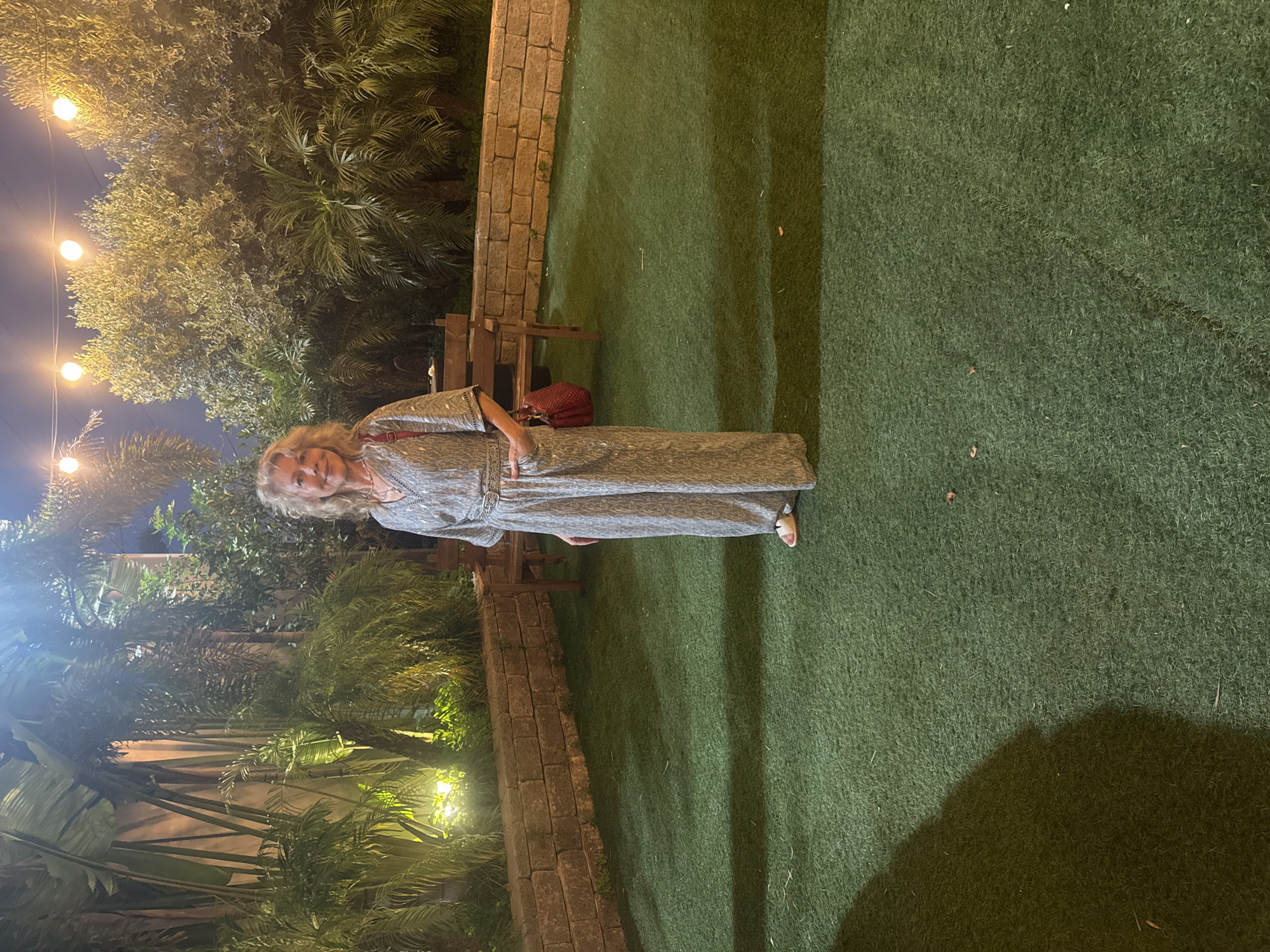 A woman with blonde, wavy hair stands in a backyard at night. She is wearing a silver patterned jumpsuit with a belt and white sandals. She has a red purse slung over her shoulder. The woman is standing on a patch of green artificial turf, with a brick wall and several benches behind her. Beyond the wall, palm trees and other greenery are visible, illuminated by overhead string lights. The scene appears to be a dimly lit outdoor seating area. The location is described as Migdal HaEmek, Israel.
