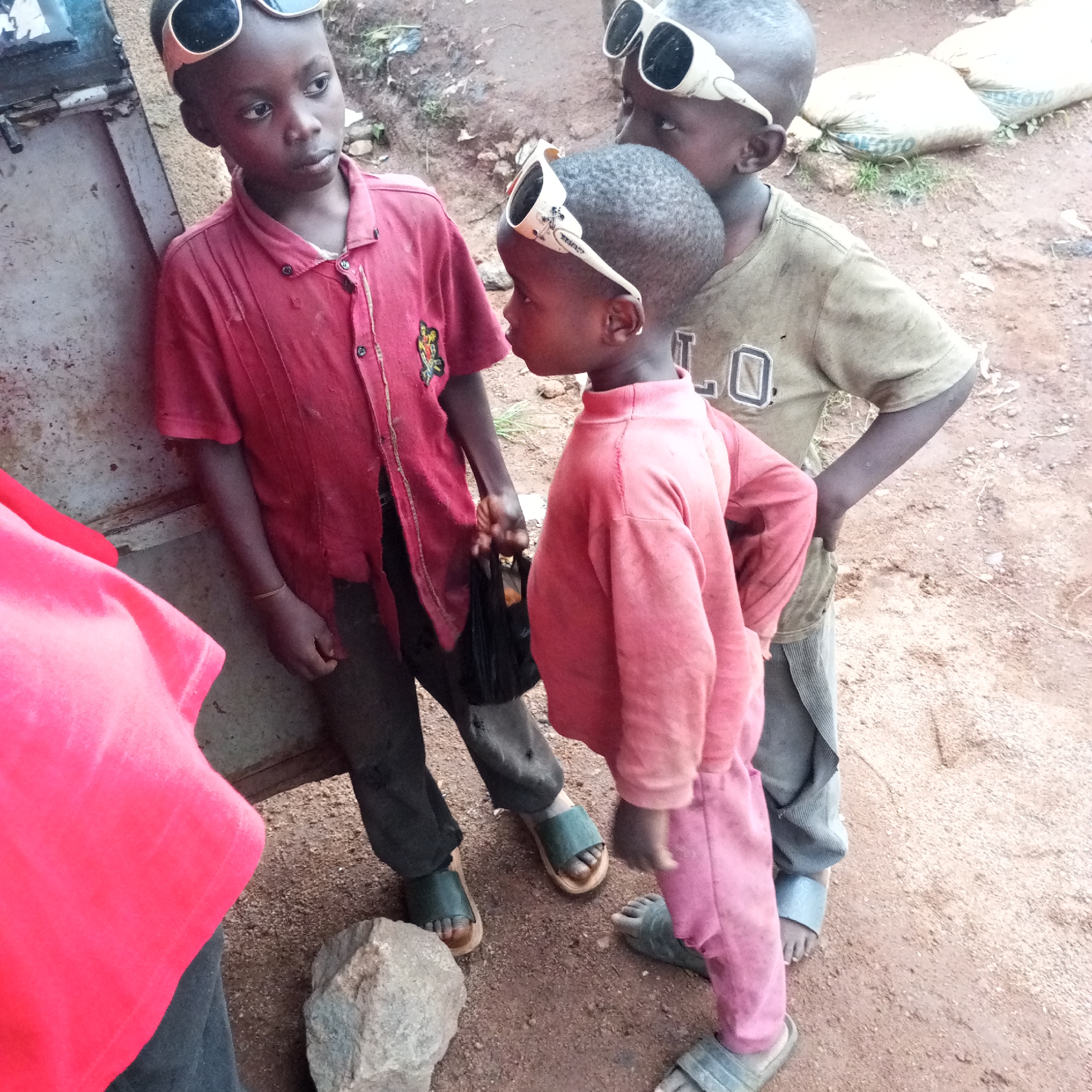 In Bukuru, Nigeria, four children are pictured outdoors. Two boys stand in the foreground, facing each other. The boy on the left wears a red collared shirt and black pants, with green slides. The boy on the right wears a pink long-sleeved shirt and pink pants, also with green slides. Both boys have white sunglasses perched on their foreheads. Behind them, another boy is visible from the chest up, wearing a tan t-shirt with "L O" logo and white sunglasses on his forehead. His skin tone is darker than the other boys. A fourth child, wearing a red shirt, stands to the far left of the frame, their face obscured. The ground is dirt and gravel, with some rocks scattered around. In the background, large sacks are stacked, possibly containing goods. The overall scene suggests a casual, everyday outdoor setting.