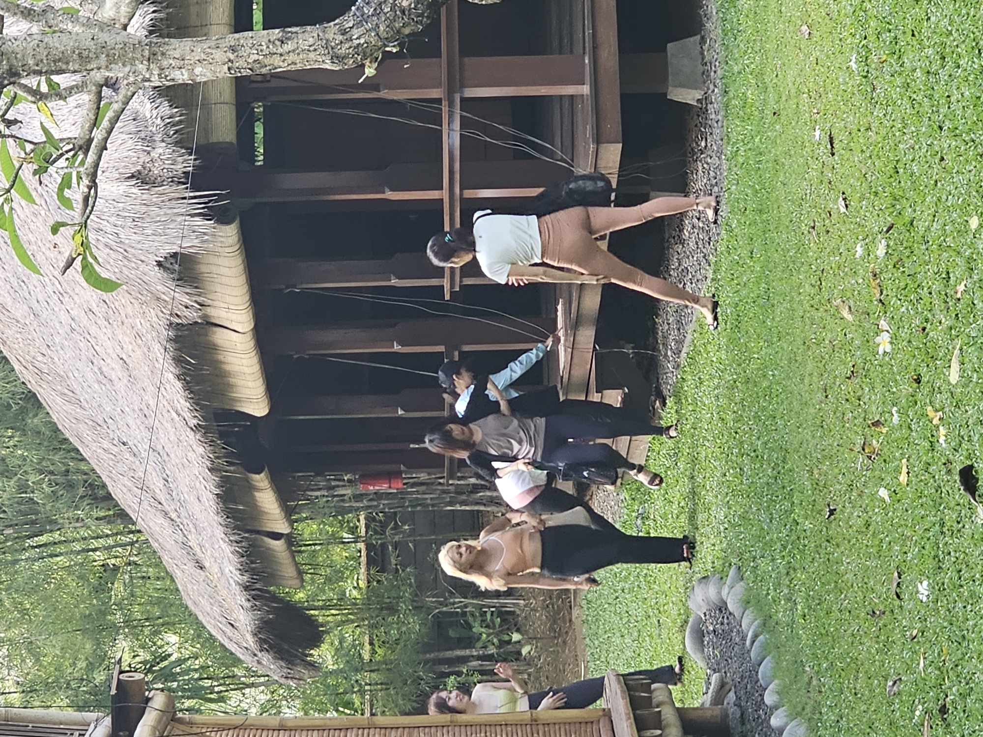 The image shows a group of women engaged in yoga or stretching exercises outdoors, beside a building with a thatched roof. The scene appears to be taking place on a grassy lawn bordered by a stone edging. The environment suggests a tropical or semi-tropical location, with lush greenery in the background. The location is Denpasar, Indonesia.