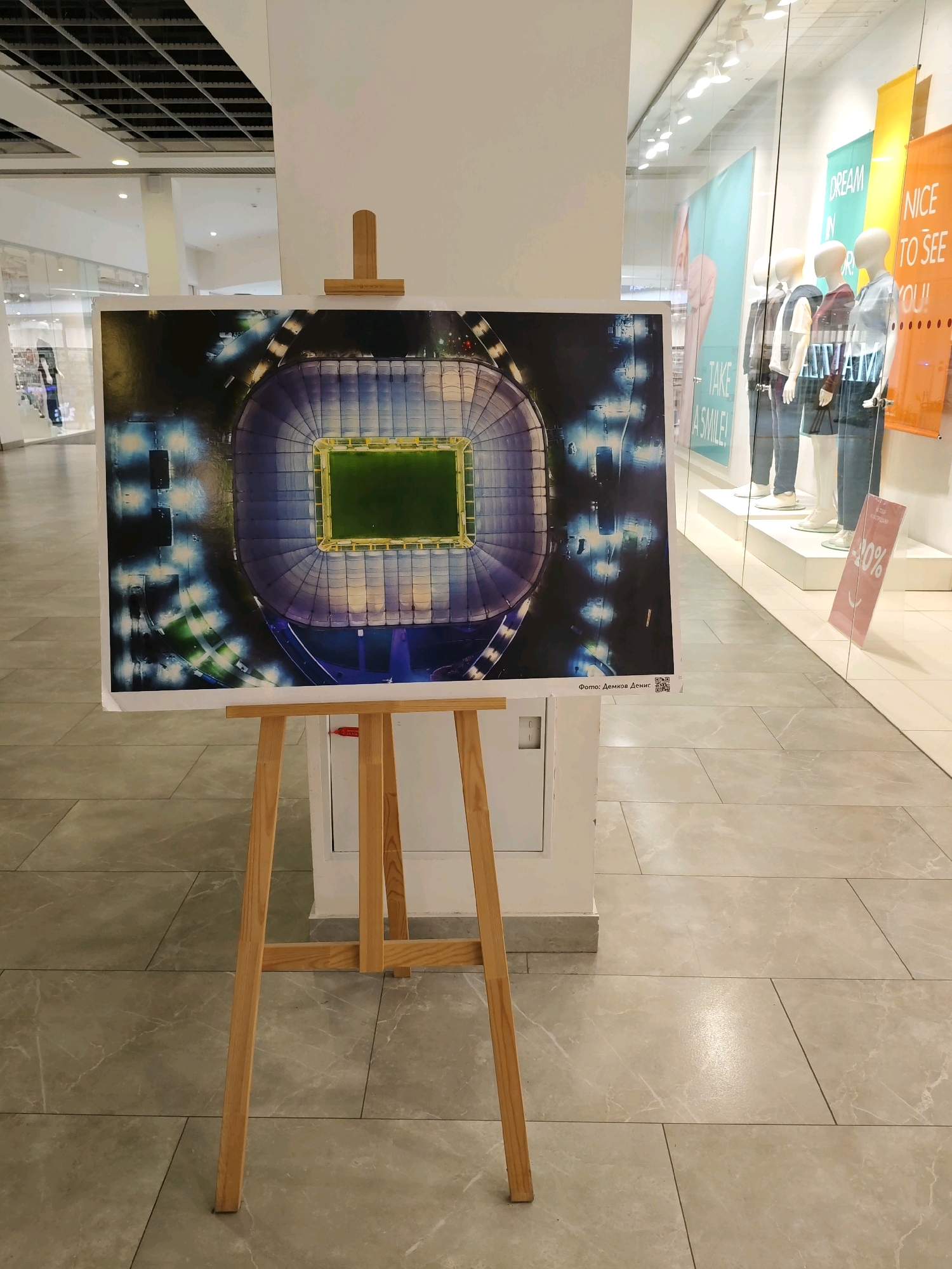 A photograph of the Rostov Arena stadium, taken from directly above, is displayed on a wooden easel in a public indoor space. The stadium, a modern football stadium with a distinctive blue and white exterior and a bright green field, is illuminated by stadium lights. The photograph is framed by a white mat. A QR code and text in Russian, "Фото: Демков Денис" (Photo: Demkov Denis), are visible in the bottom right corner of the photograph. The background of the image reveals a retail environment with mannequins in a shop window displaying clothing. One banner behind the mannequins reads "NICE TO SEE YOU!". The floor is tiled with a light marble pattern. The location is the Rostov Arena in Rostov-on-Don, Russia, on display in what appears to be a shopping mall.
