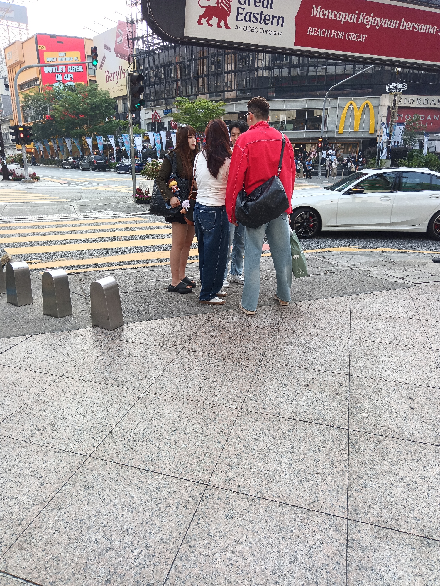 The image captures a street scene in Kuala Lumpur, Malaysia, likely in the bustling Bukit Bintang area, characterized by its prominent advertising billboards and commercial establishments. In the foreground, four individuals are standing on a granite-tiled sidewalk at a pedestrian crossing. Two women in casual attire, one with dark hair and a jacket, the other with lighter hair and a white top, are conversing. They are accompanied by two men, one wearing a bright red jacket and a dark crossbody bag, and another partially obscured man in a light-colored t-shirt. A white sedan is visible in the background, stopped at the intersection, with McDonald's and Giordano storefronts further down the street. Traffic lights and street signs are also present, indicating a busy urban environment. The overall scene depicts a moment of pause amidst the city's activity, with pedestrians waiting or interacting at a crosswalk.
