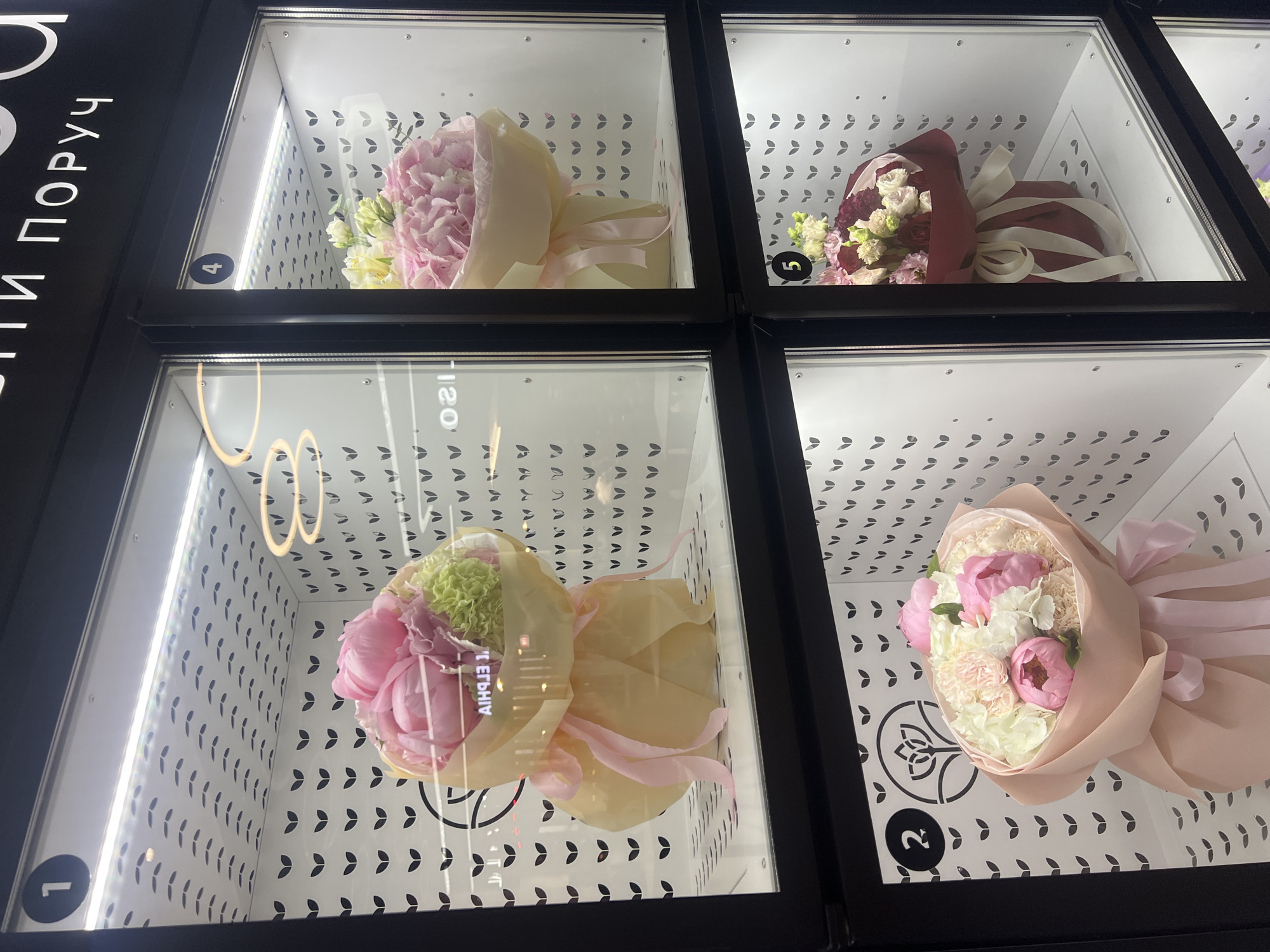 This image captures a close-up view of a modern flower vending machine or refrigerated display case, likely situated indoors in Kyiv, Ukraine. The machine features a black frame with multiple glass-fronted compartments, four of which are visible and numbered 1, 2, 4, and 5. Each compartment is brightly illuminated from within, showcasing a carefully arranged bouquet of flowers. The interior walls of the display units are white with a repeating pattern of small, black, stylized leaf shapes.
The four visible bouquets are:
* **Compartment 4 (top left):** A bouquet of predominantly light pink large flowers (likely peonies or hydrangeas) mixed with some smaller white/cream blossoms, wrapped in light cream-colored paper and tied with a pale pink ribbon.
* **Compartment 5 (top right):** A bouquet featuring dark red and white smaller flowers (possibly roses or carnations), wrapped in a deep burgundy paper and secured with a cream-colored ribbon.
* **Compartment 1 (bottom left):** A bouquet with prominent pink large flowers (peonies) and bright green, frilly florets, encased in light yellow wrapping paper with pink ribbons.
* **Compartment 2 (bottom right):** A mixed bouquet of pink large flowers (peonies/carnations) and smaller white blossoms, wrapped in a light peach-colored paper and tied with a pink ribbon.
On the left side of the machine, vertical Cyrillic text is partially visible, reading "И ПОРУЧ", which likely translates to "and order" or "to order", suggesting a self-service or customization option. A stylized floral or leaf logo is also visible within compartments 1 and 2. Reflections on the glass fronts distort some interior details and show reversed text, like "12O". The overall setting appears clean, commercial, and designed to present the flowers appealingly for purchase.