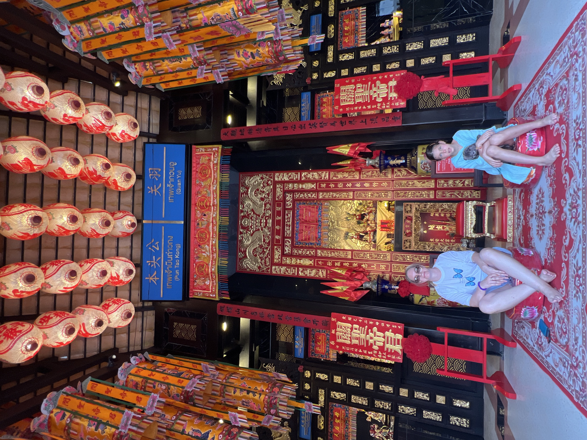 This is a full-body, front-facing photograph of two individuals, a woman and a young girl, posing in a brightly decorated temple in Baan Hua Thanon, Thailand. The temple interior is opulent, with red and gold dominant colors. Elaborate Chinese lanterns hang from the ceiling, and ornate banners with Chinese calligraphy adorn the walls. In the background, a shrine is visible, featuring golden statues and red fabric.
The woman, on the left, is seated on a red cylindrical cushion, wearing sunglasses, a light blue t-shirt with butterfly prints, and light-colored shorts. Her legs are crossed, and her arms are resting on her lap.
The young girl, on the right, is also seated on a similar red cushion, holding a small gray stuffed animal. She is wearing a light blue, loose-fitting garment and has her legs crossed.
Both individuals are positioned on a large, red carpet with intricate white patterns, placed on a tiled floor. Red wooden chairs and stands are strategically placed around the scene, enhancing the temple's traditional aesthetic. The overall impression is one of a cultural visit, with the subjects engaging with the visually rich environment.